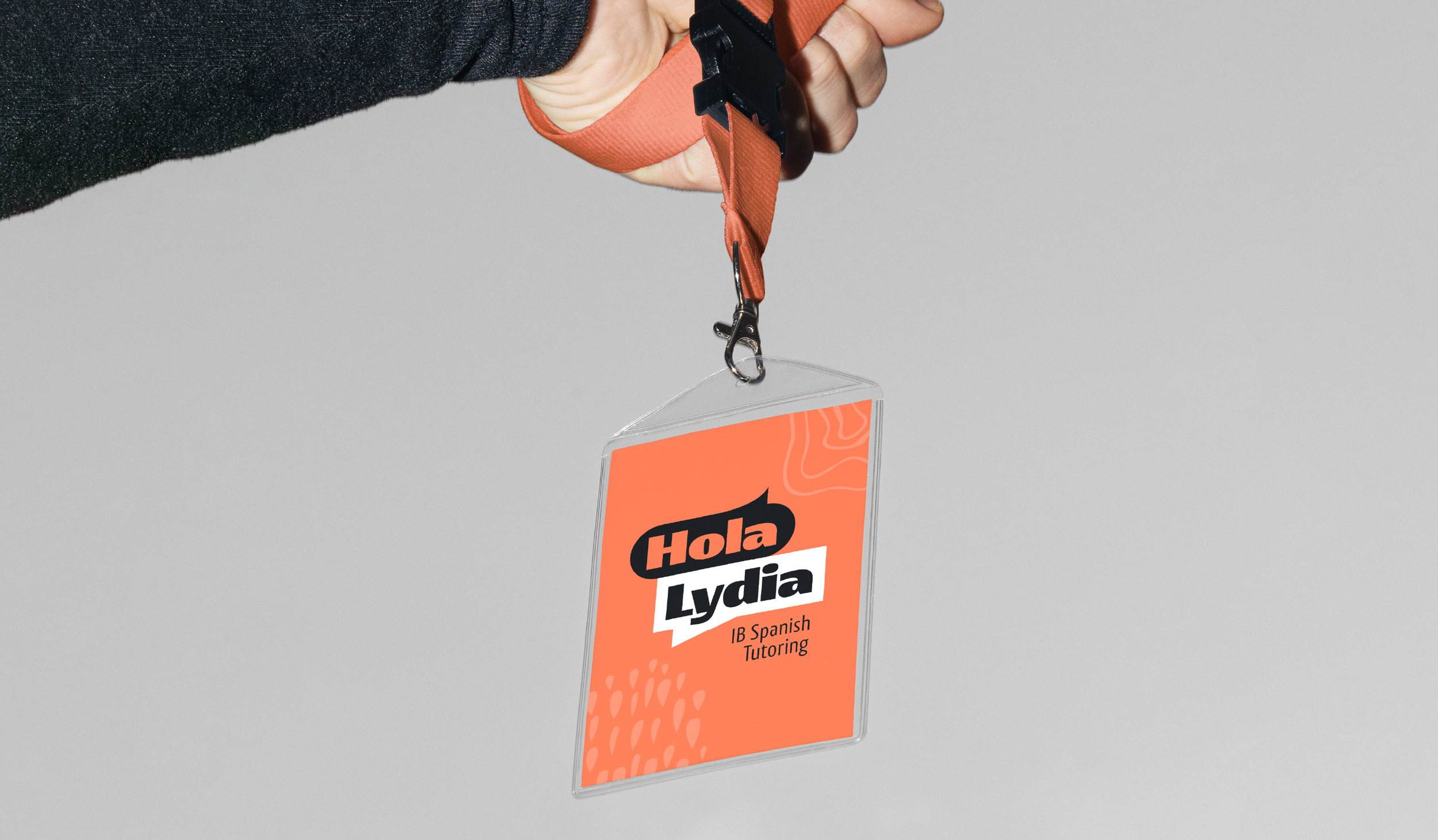 Close-up of a person's hand holding a black clip attached to an orange lanyard with a clear plastic badge holder displaying a colorful card that reads 'Hola Lydia,' indicating IB Spanish tutoring.