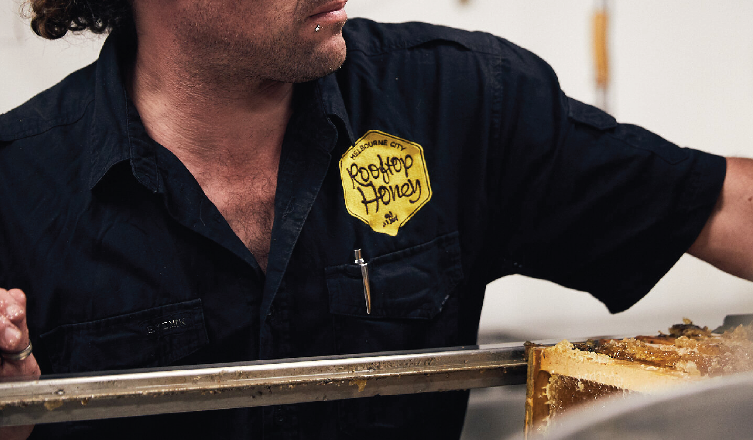 A person wearing a navy blue uniform with a yellow patch that reads 'Melbourne City Rooftop Honey' is standing behind a tray of honeycomb, observing or preparing the honeycomb.