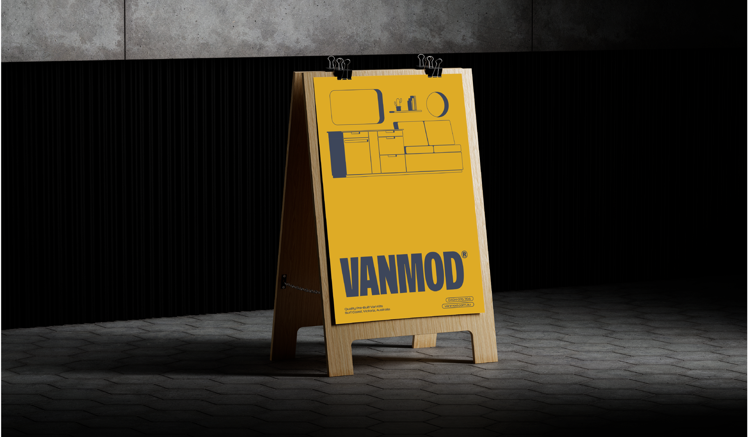 A yellow signboard with the text 'VANMOD' leaning against a wooden frame, placed on a dark tiled floor against a black wall.