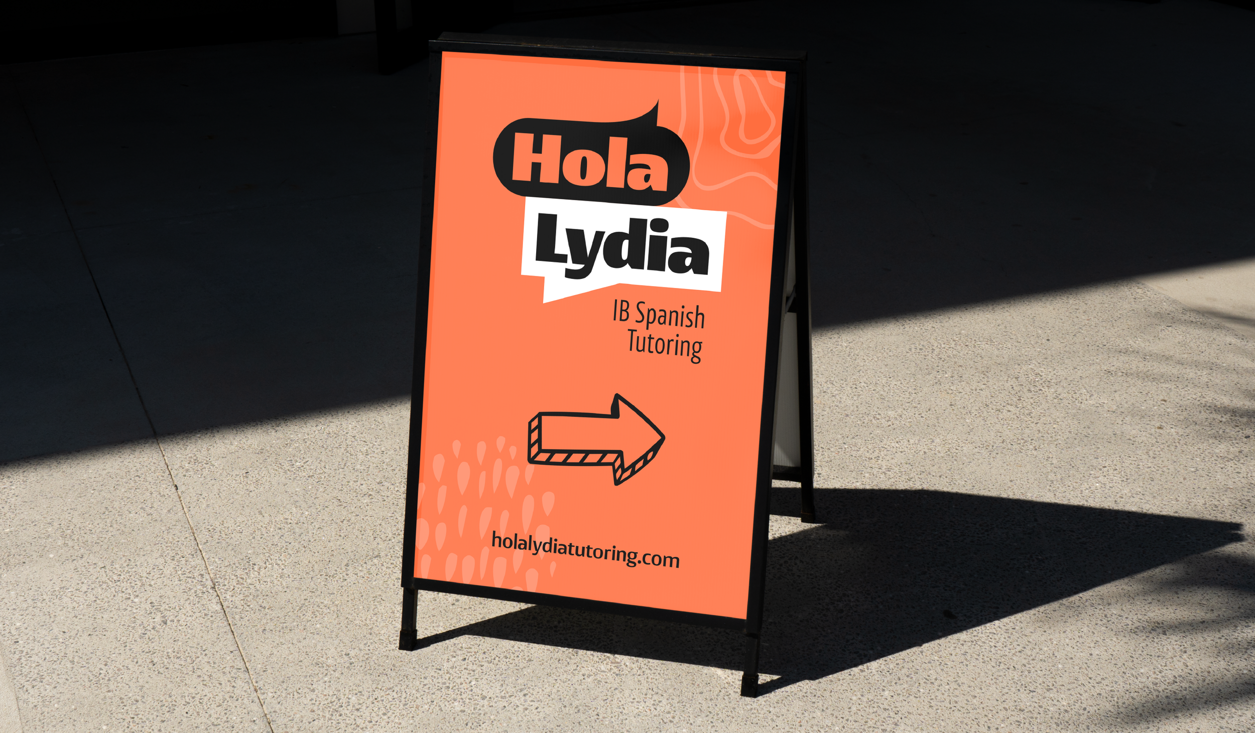 Bright orange sandwich board sign with black and white text that reads "Hola Lydia" and "IB Spanish Tutoring," with a rightward arrow at the bottom. The sign is on a sidewalk with shadows in the background.