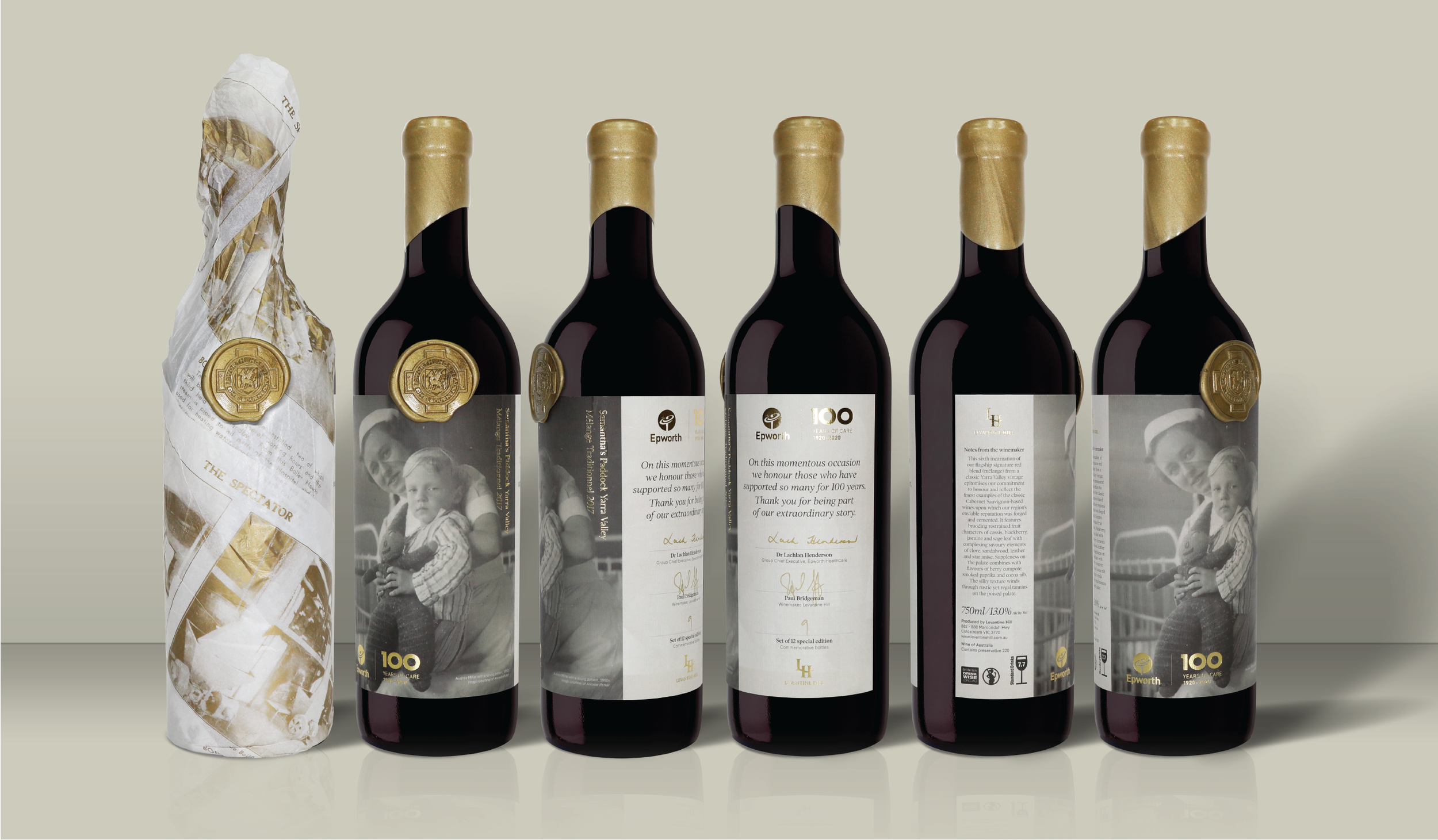 A row of six wine bottles with black labels and gold foil caps, five of which have custom labels featuring black-and-white images of a girl with a dog, while the first bottle is wrapped in decorative paper with gold seals.