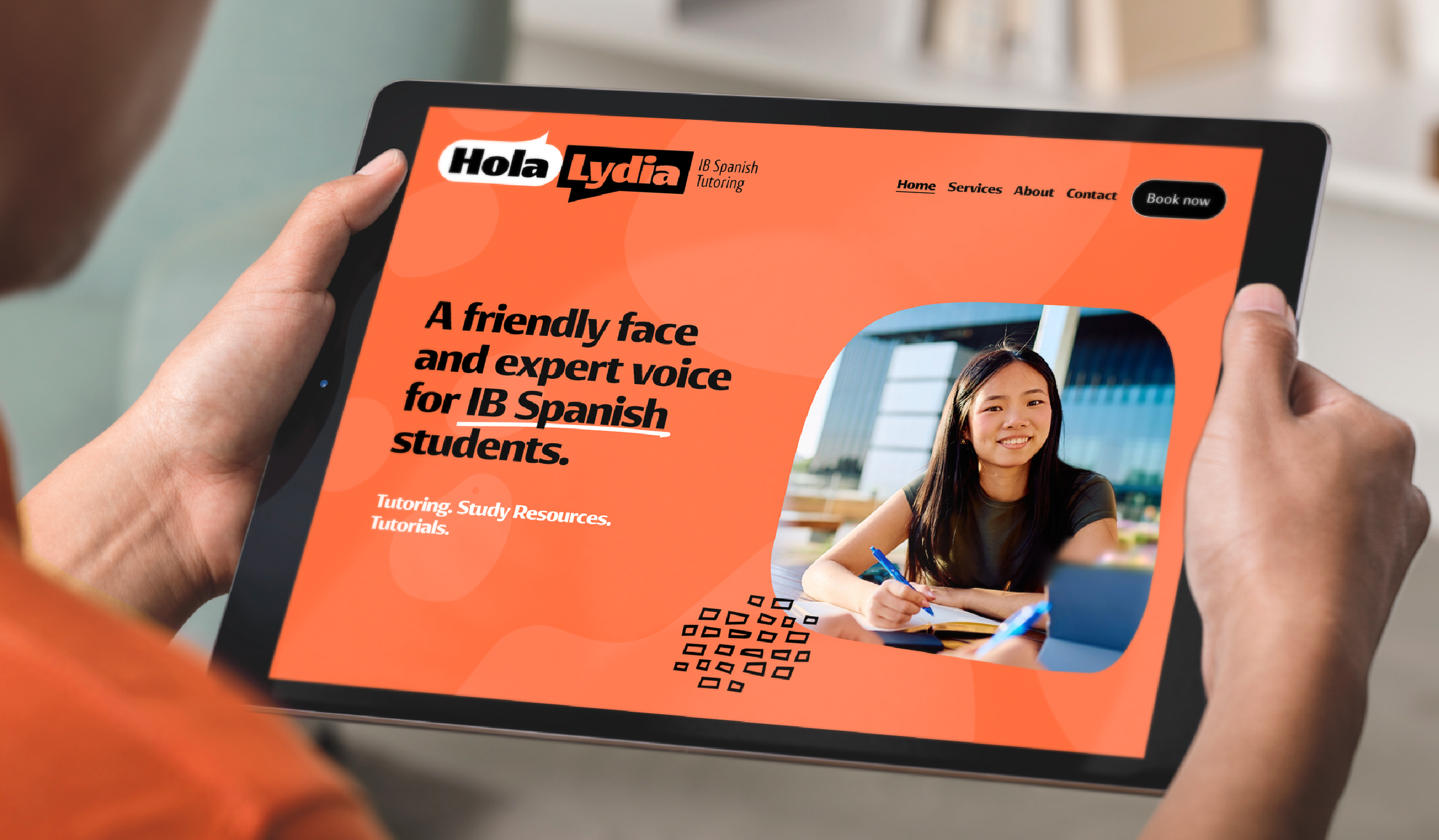 A person holding a tablet displaying a website for Lydia, an IB Spanish tutoring service. The webpage features a logo with the text "Hola Lydia" and has a menu with links to Home, Services, About, Contact, and a black "Book now" button. The main text on the page says, "A friendly face and expert voice for IB Spanish students," with accompanying image of a smiling young woman writing with a blue pen at a desk.