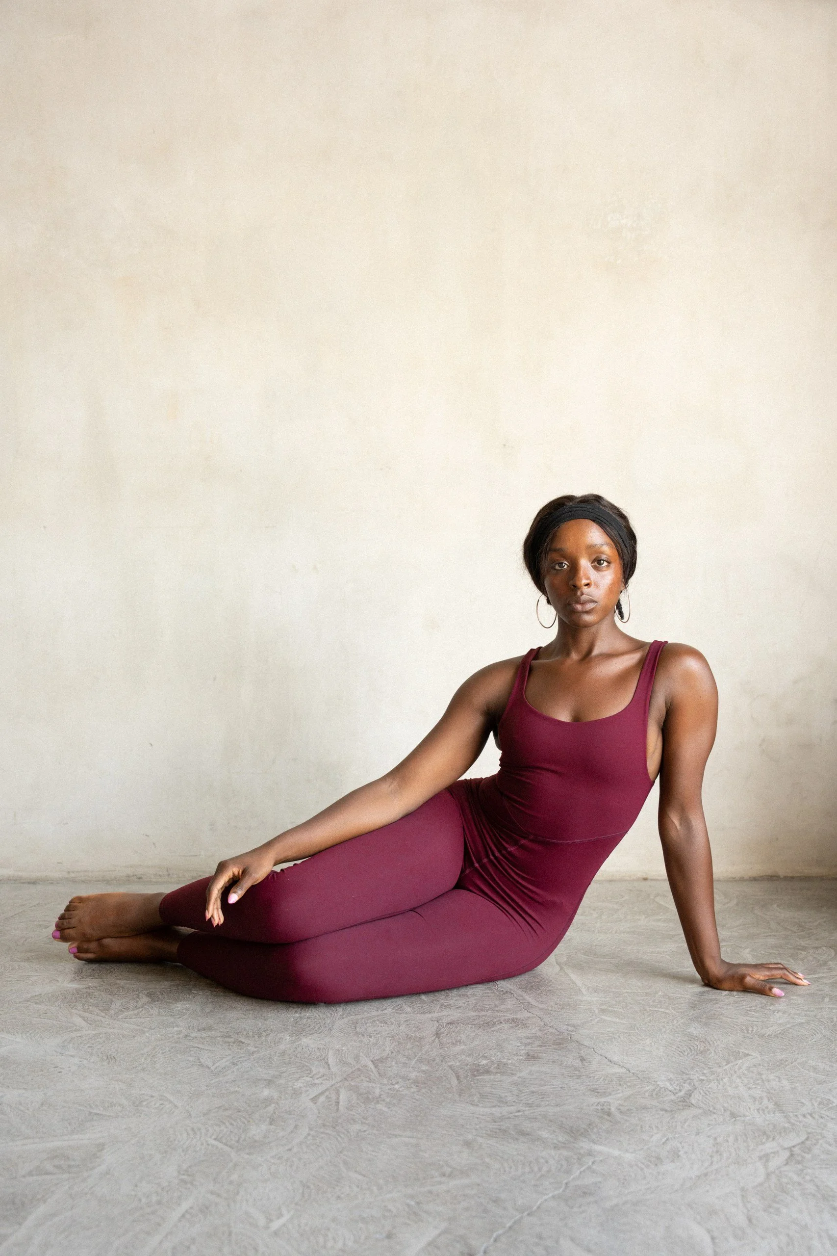 A woman in a maroon sleeveless bodysuit sitting on the floor in a side pose, with her legs extended and one arm resting on the ground, against a plain off-white wall.