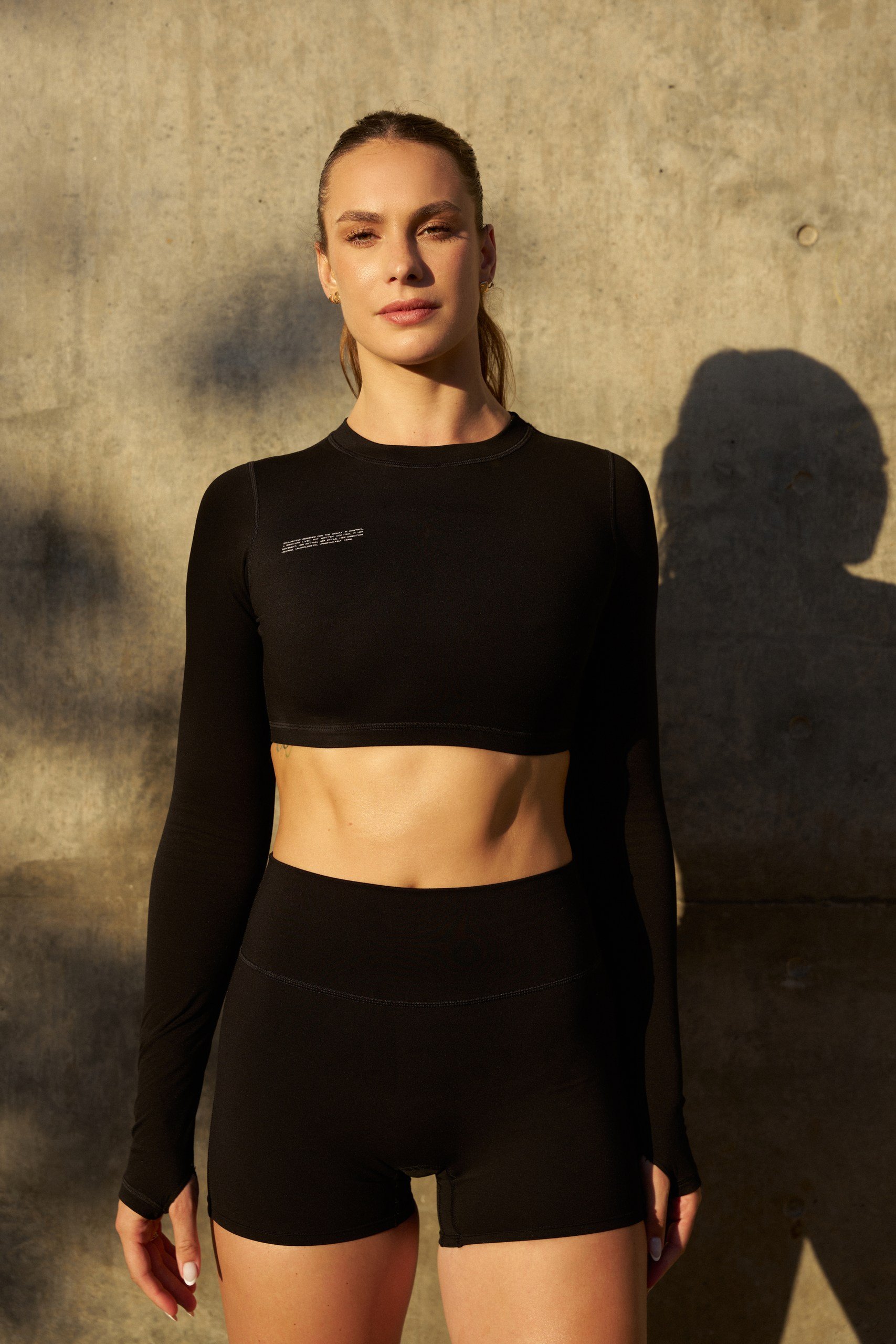 A woman standing against a concrete wall, wearing a black long-sleeve athletic crop top and black shorts, with her shadow cast on the wall behind her.