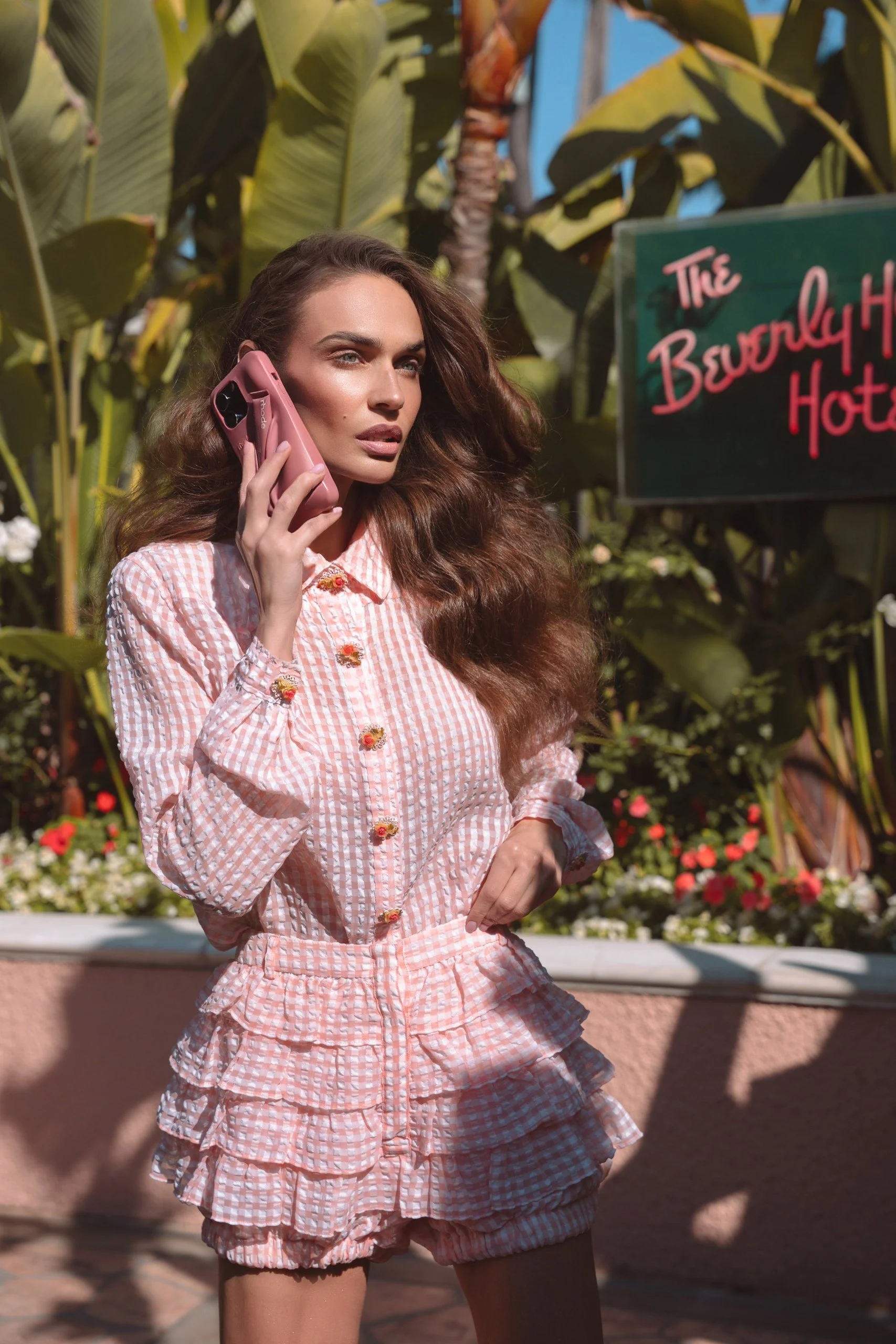 A woman with long wavy brown hair is talking on a pink phone and standing outdoors in front of green tropical plants and a sign that reads 'The Beverly Hills Hotel'.