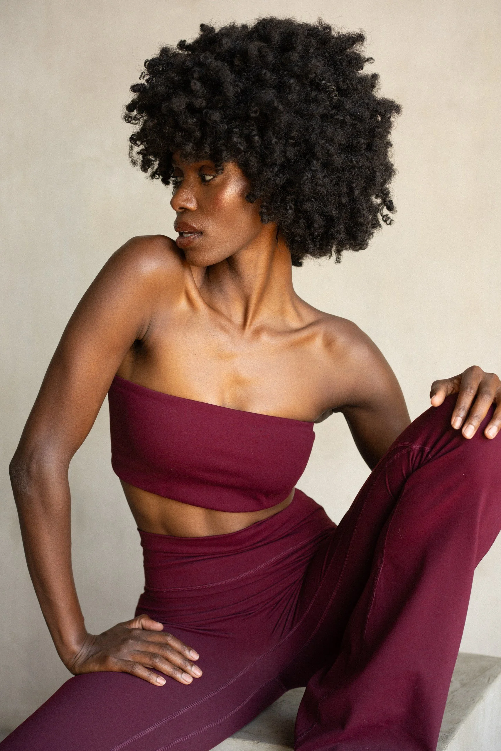 A woman with dark skin and natural curly hair in a short style, wearing a maroon sports bra and matching leggings, poses against a plain background.