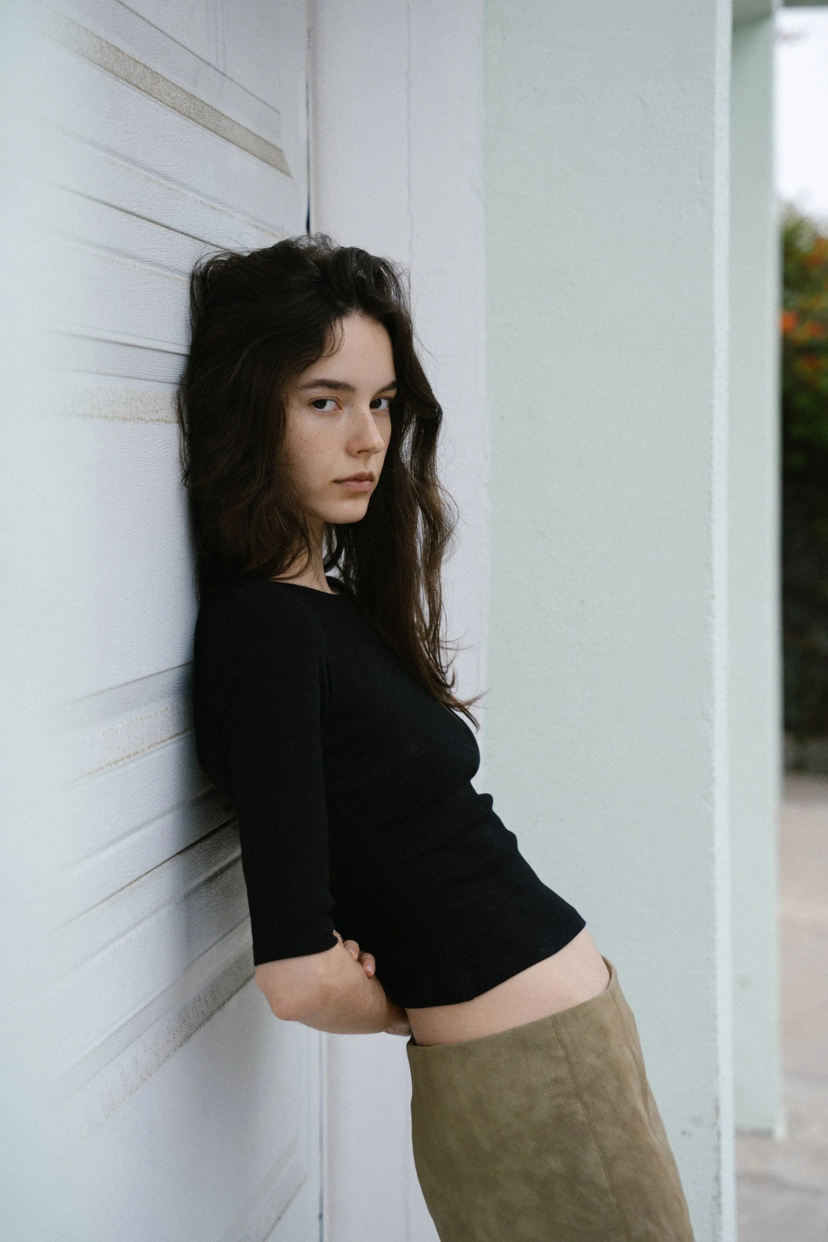 A young woman with long dark hair wears a black top and beige skirt, leaning against a white wooden wall outside.