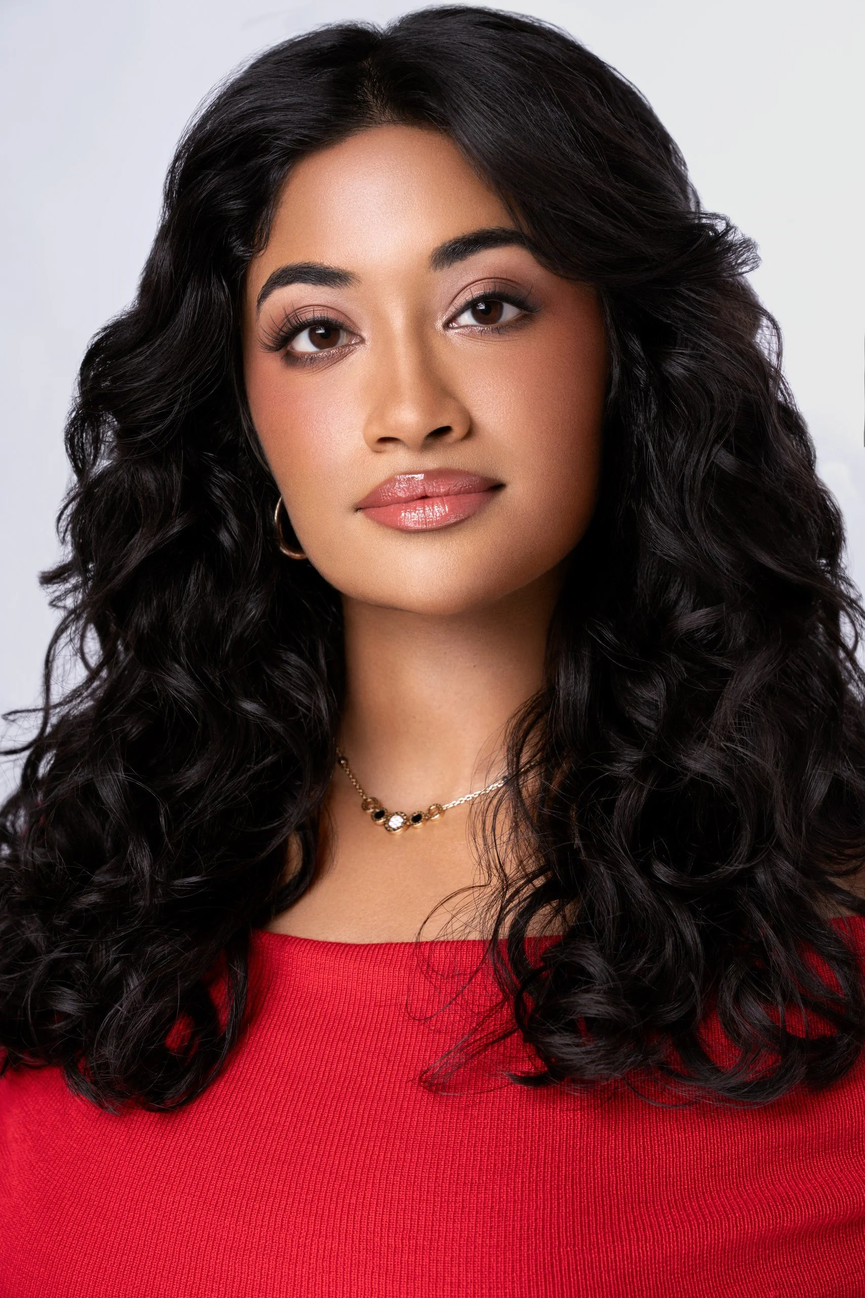 Close-up of a woman with long, curly black hair, wearing makeup with defined eyebrows and glossy lips, a gold necklace, and a red top against a neutral background.