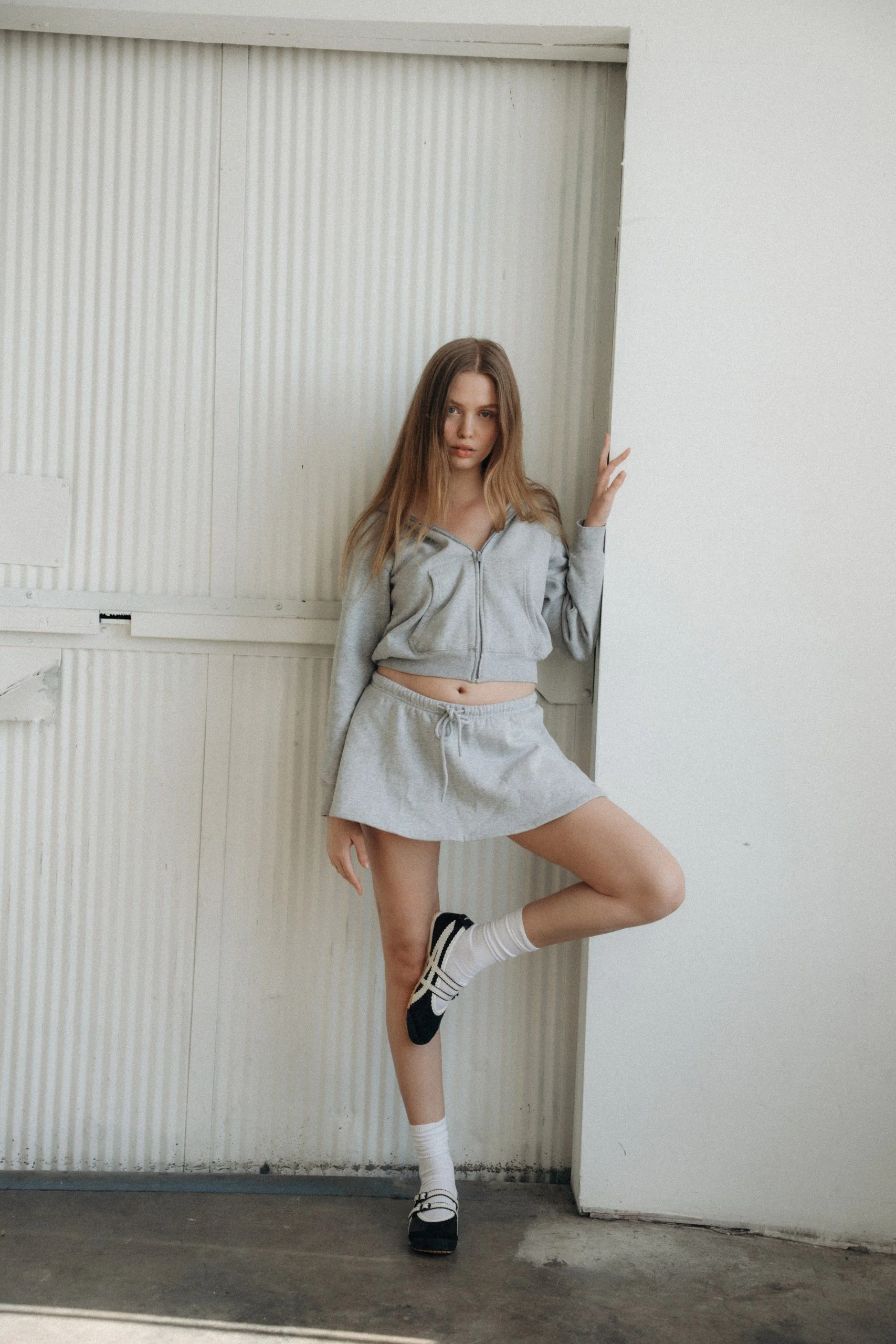 Young woman wearing a gray zip-up hoodie, matching gray shorts, and black-and-white sneakers poses with one leg raised and foot resting on the wall in a minimalist indoor setting with white walls and a corrugated wood door.