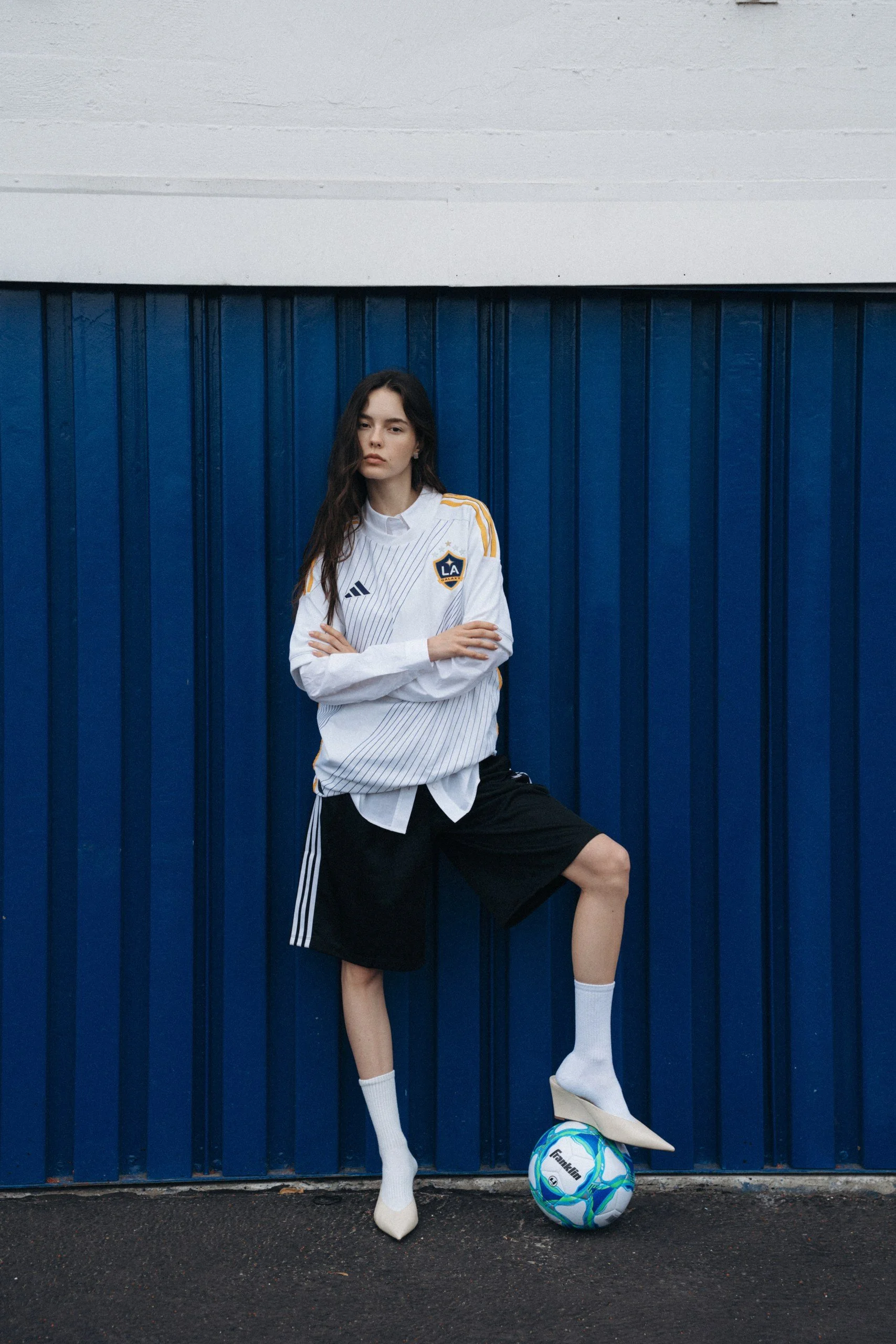 A young woman with long dark hair standing against a blue steel fence, wearing a white LA Galaxy sports jersey, black shorts, white socks, and white shoes, with one foot resting on a colorful soccer ball.