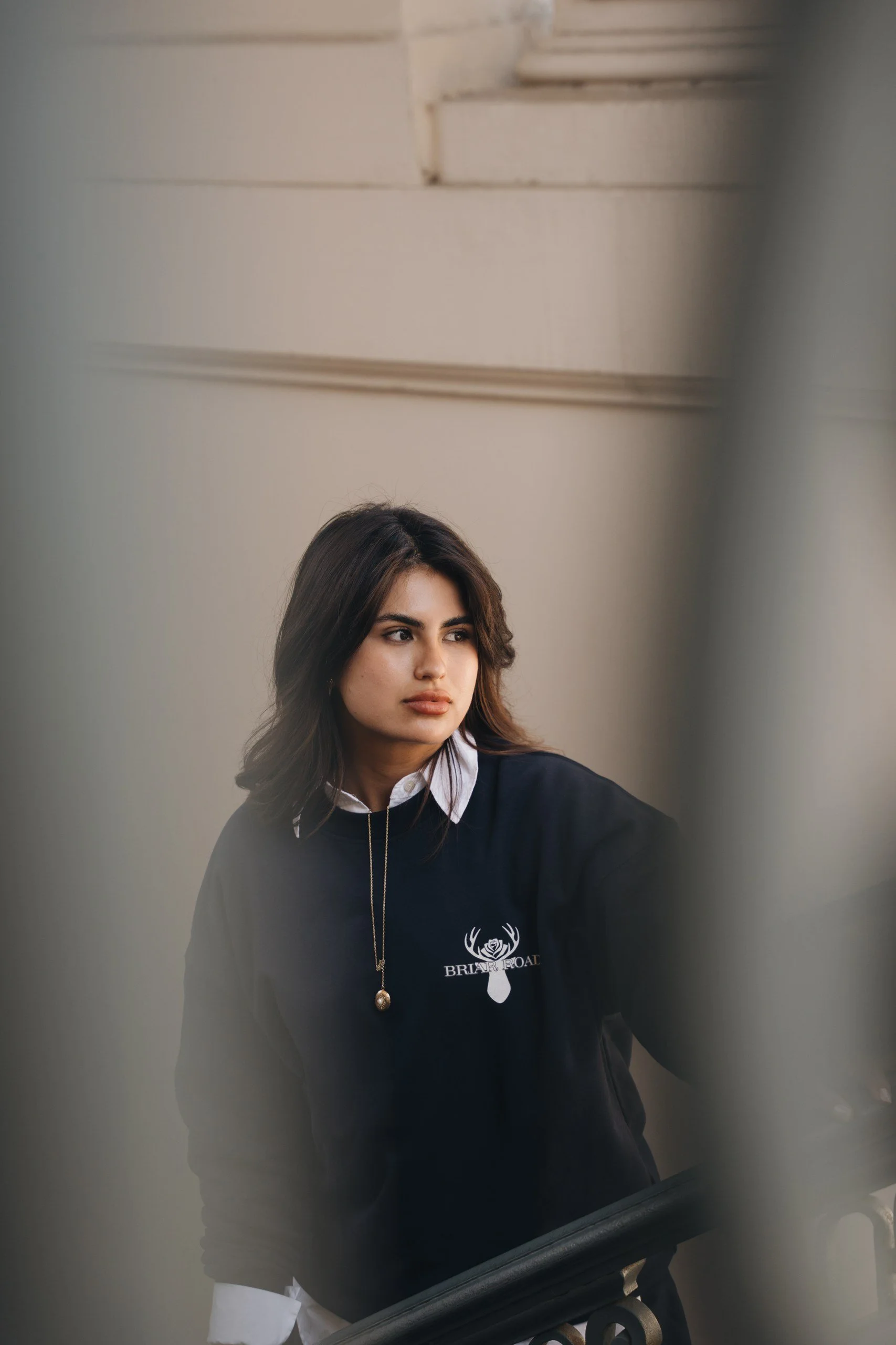 A young woman with dark hair and a serious expression, standing near a staircase, wearing a black hoodie with a logo on it and a white shirt underneath. She has a long necklace with a pendant.