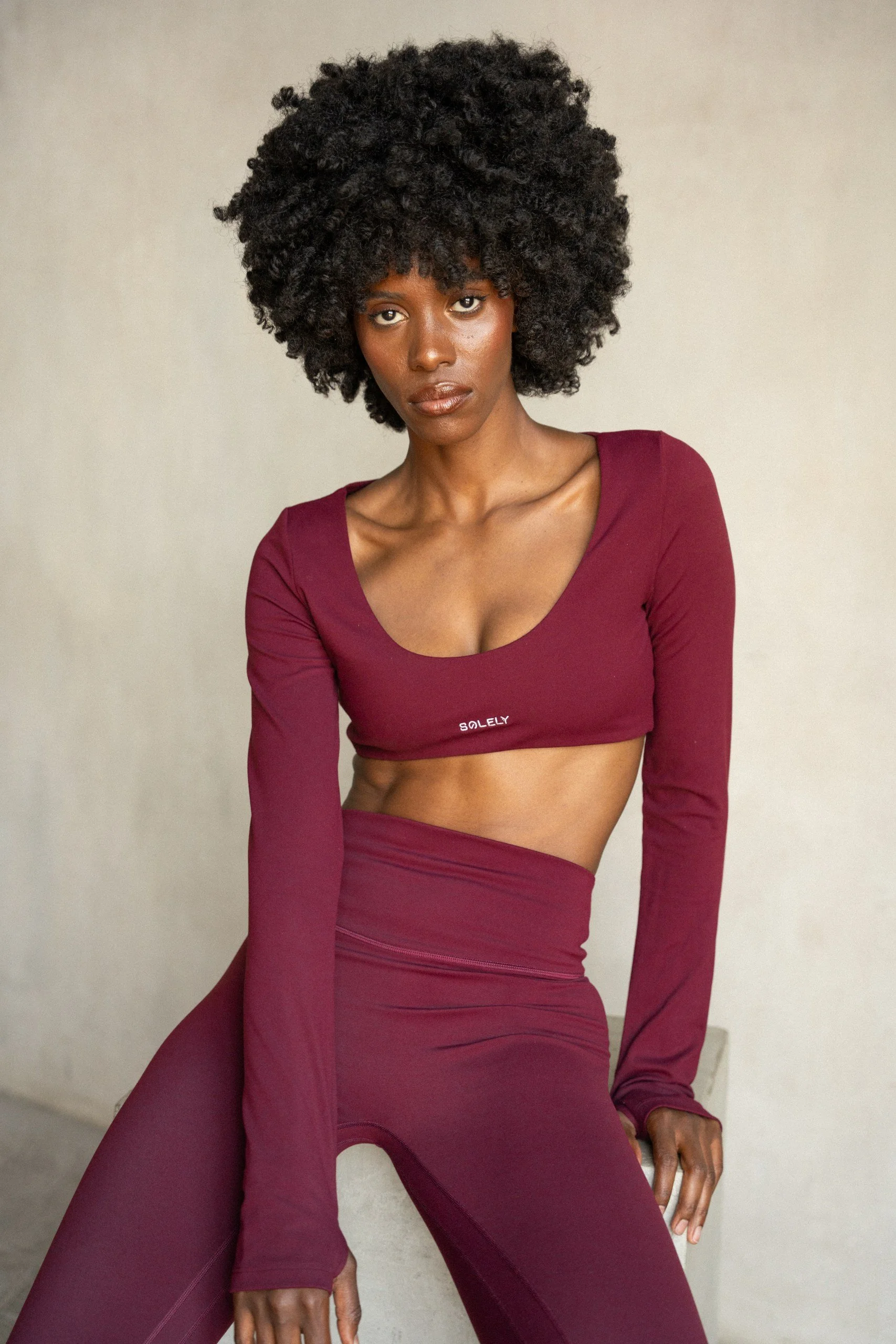 A woman with dark skin and voluminous curly black hair sitting against a plain neutral-colored wall, wearing a matching burgundy long-sleeve crop top and high-waisted leggings.
