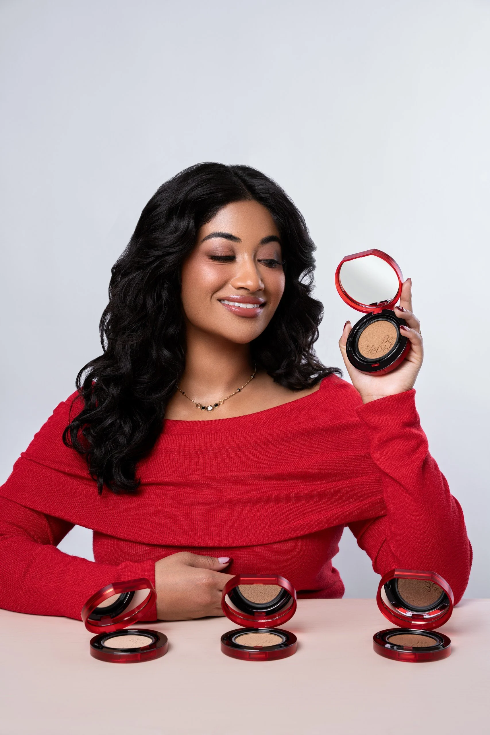 A woman in a red sweater holding a compact powder mirror with a smile, surrounded by open compact powder cases on a light surface with a plain background.