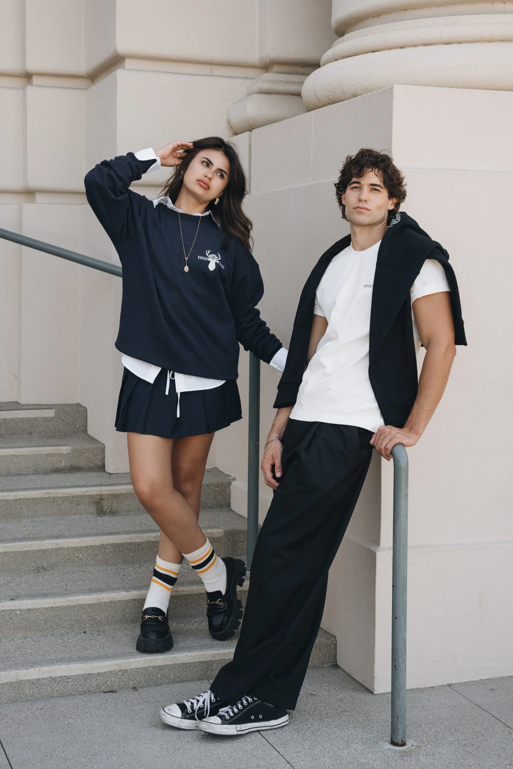 Two young adults, a woman and a man, posing outdoors on concrete stairs near a building. The woman wears a navy sweatshirt, a white collared shirt, a black skirt, and striped socks, with her hand on her head. The man wears a white t-shirt, black pant