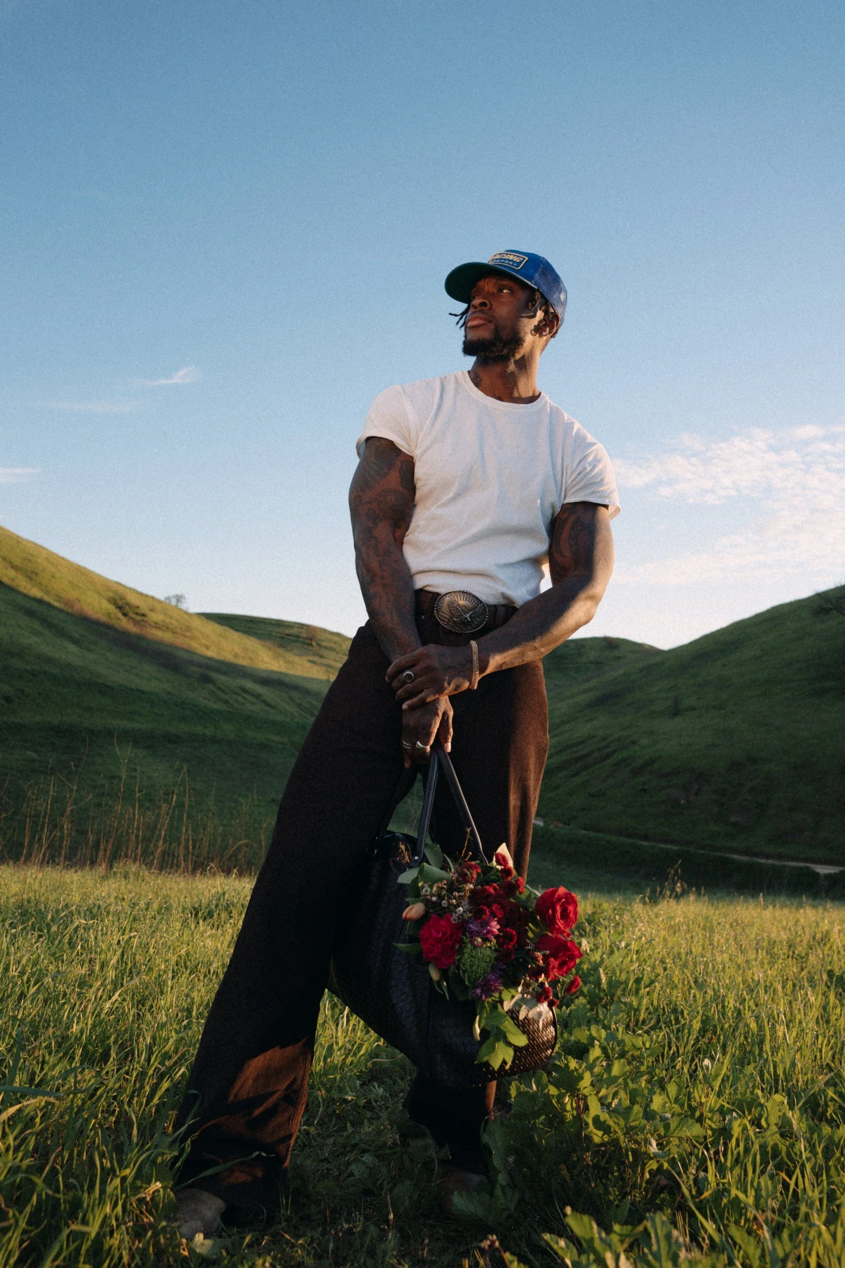 A man in a white t-shirt and dark pants stands outdoors in a green grassy field, holding a bouquet of red and purple flowers, with rolling green hills in the background under a blue sky.