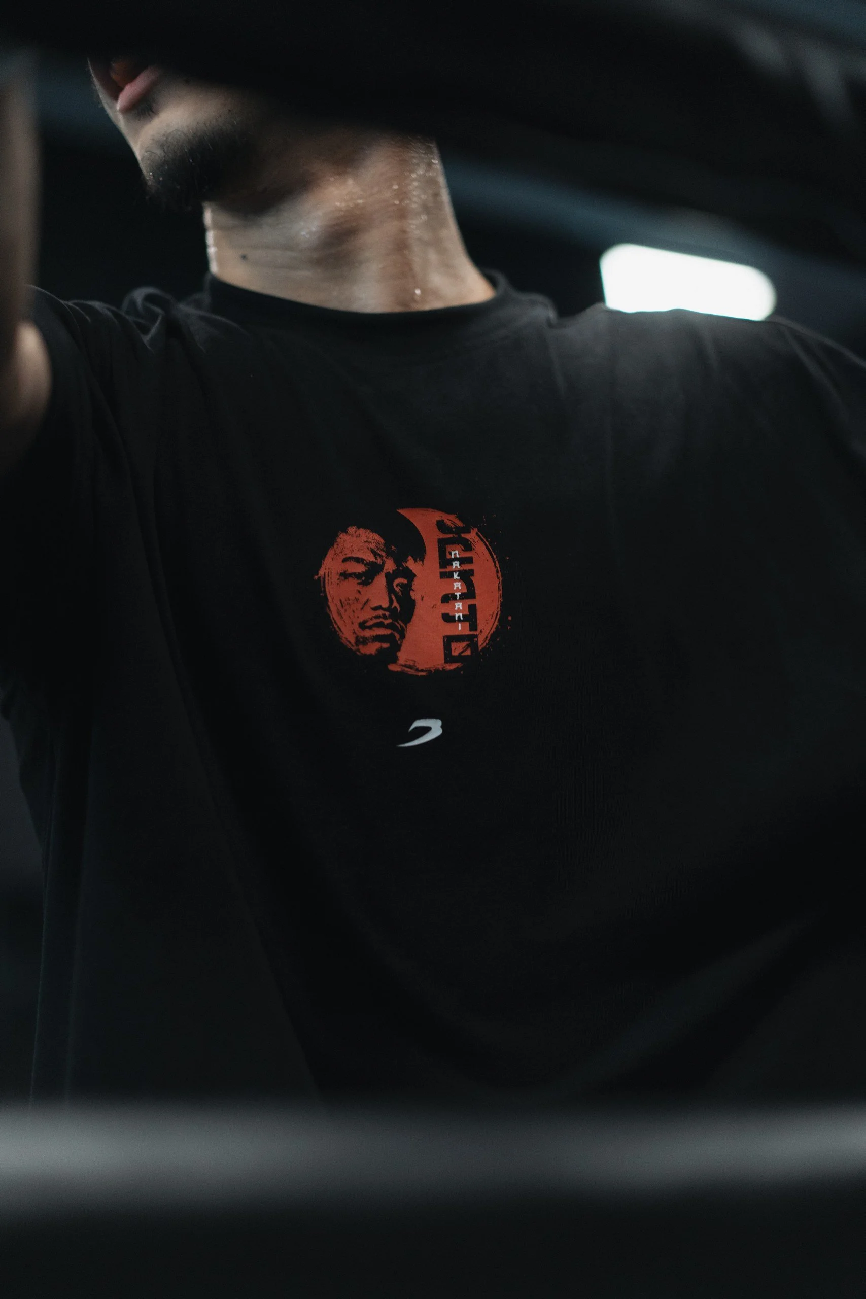 Close-up of a man wearing a black t-shirt with a red graphic and text, with his head slightly turned downward, located in a dimly lit indoor environment.