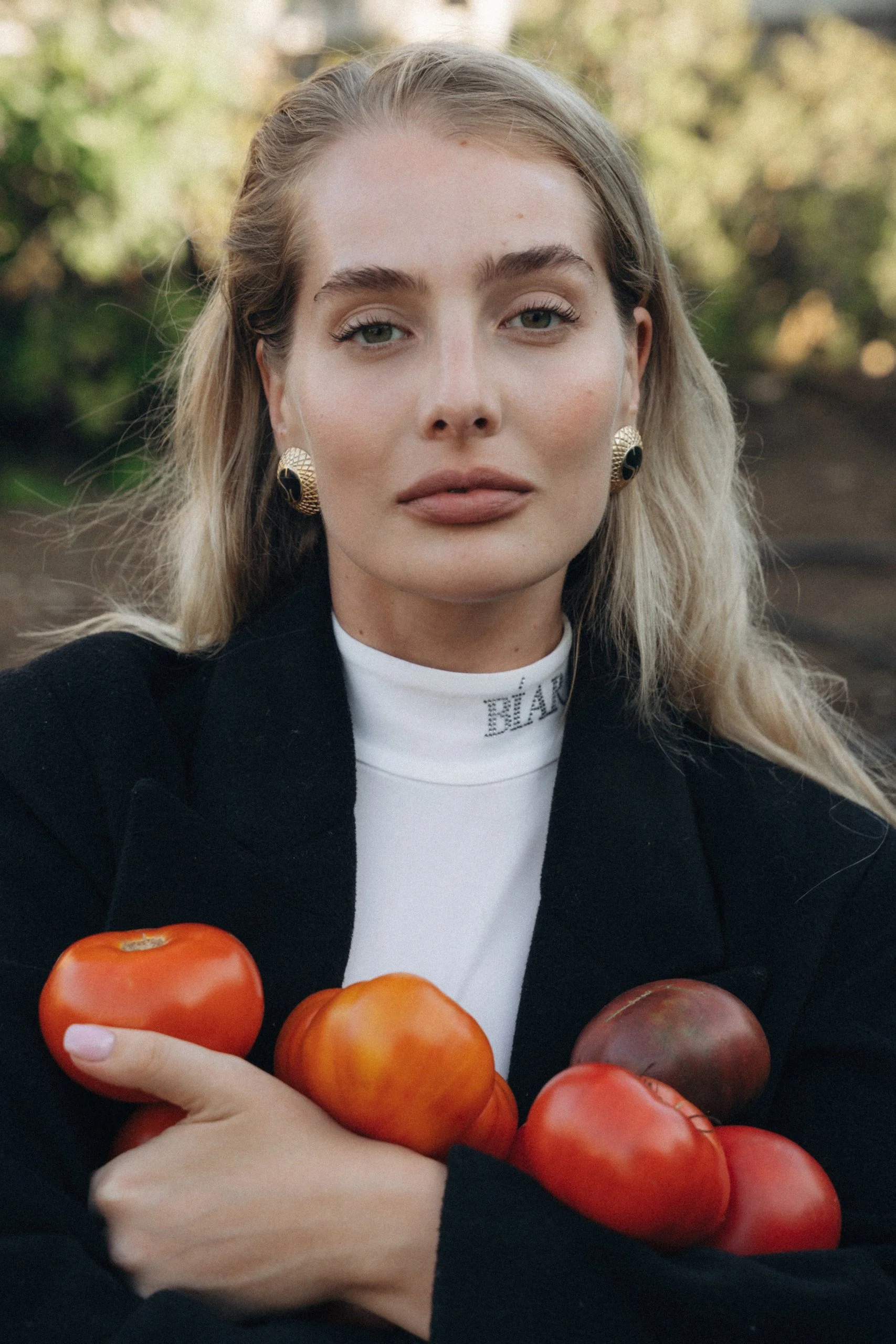 A woman with blonde hair wearing black and white clothing and gold earrings holds tomatoes and peaches in her arms outside.