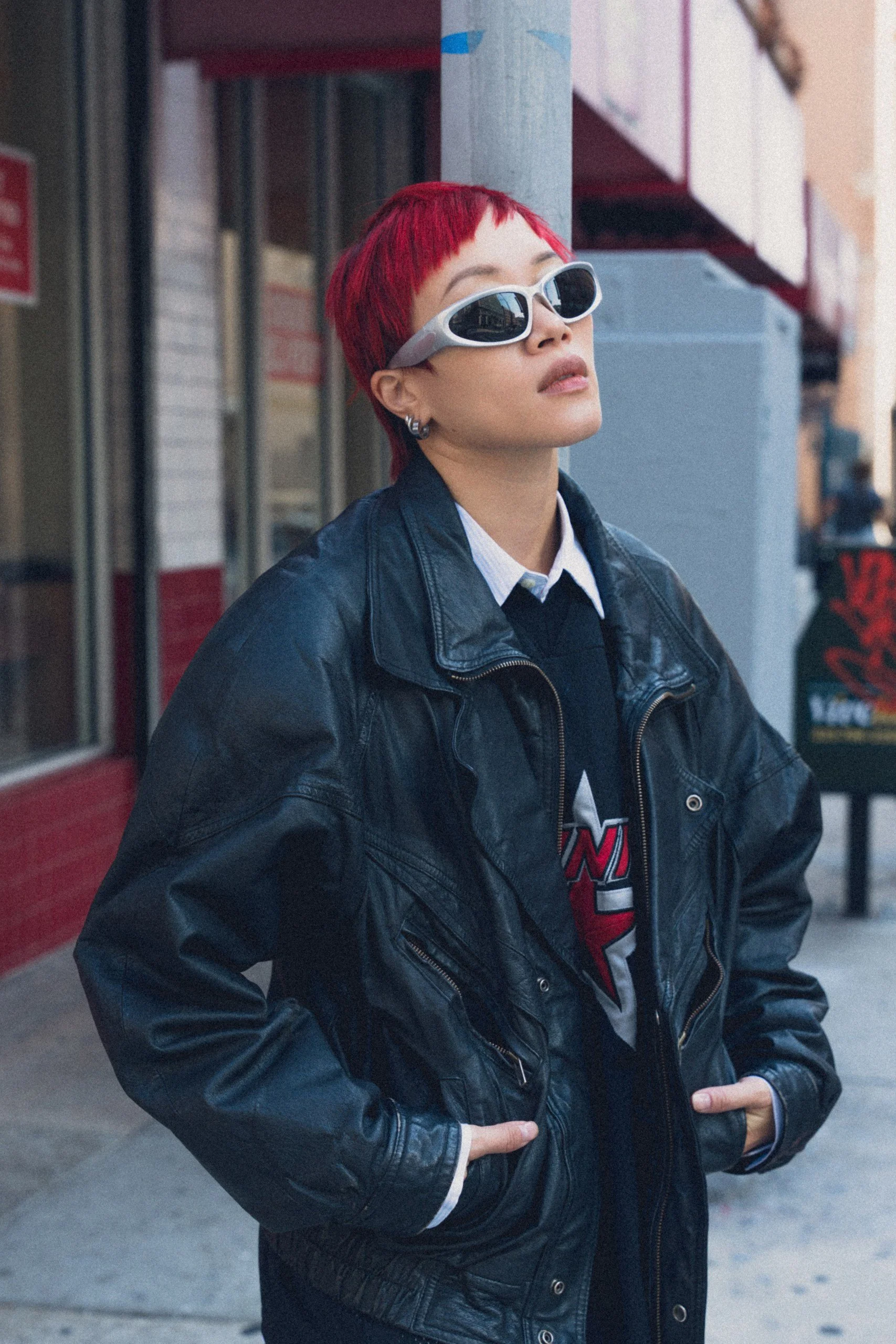 Woman with red hair wearing sunglasses and a leather jacket standing outside on a city street.