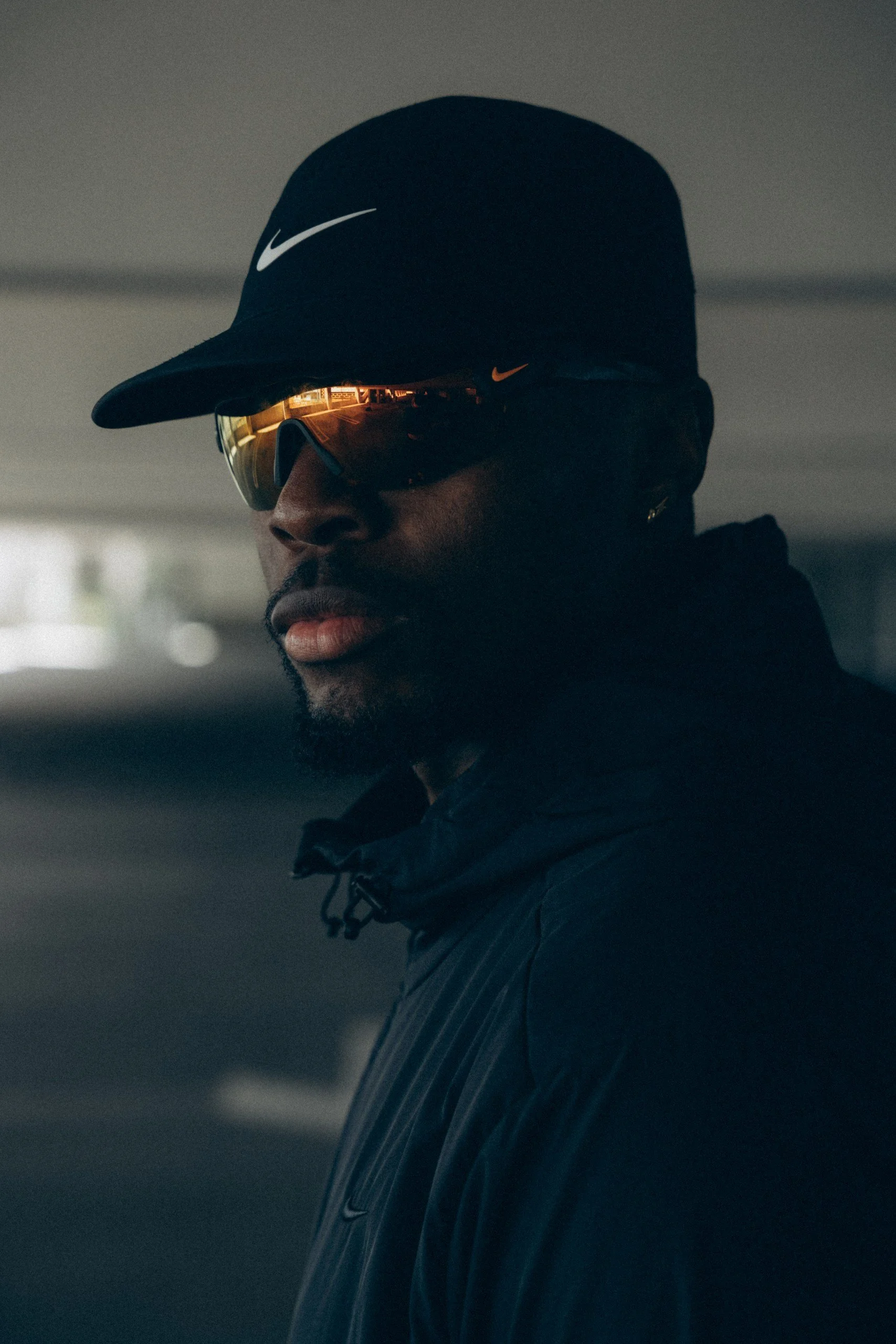 A close-up photo of a man wearing a black Nike cap, sporty sunglasses with reflective lenses, and a dark jacket, with a blurred background.