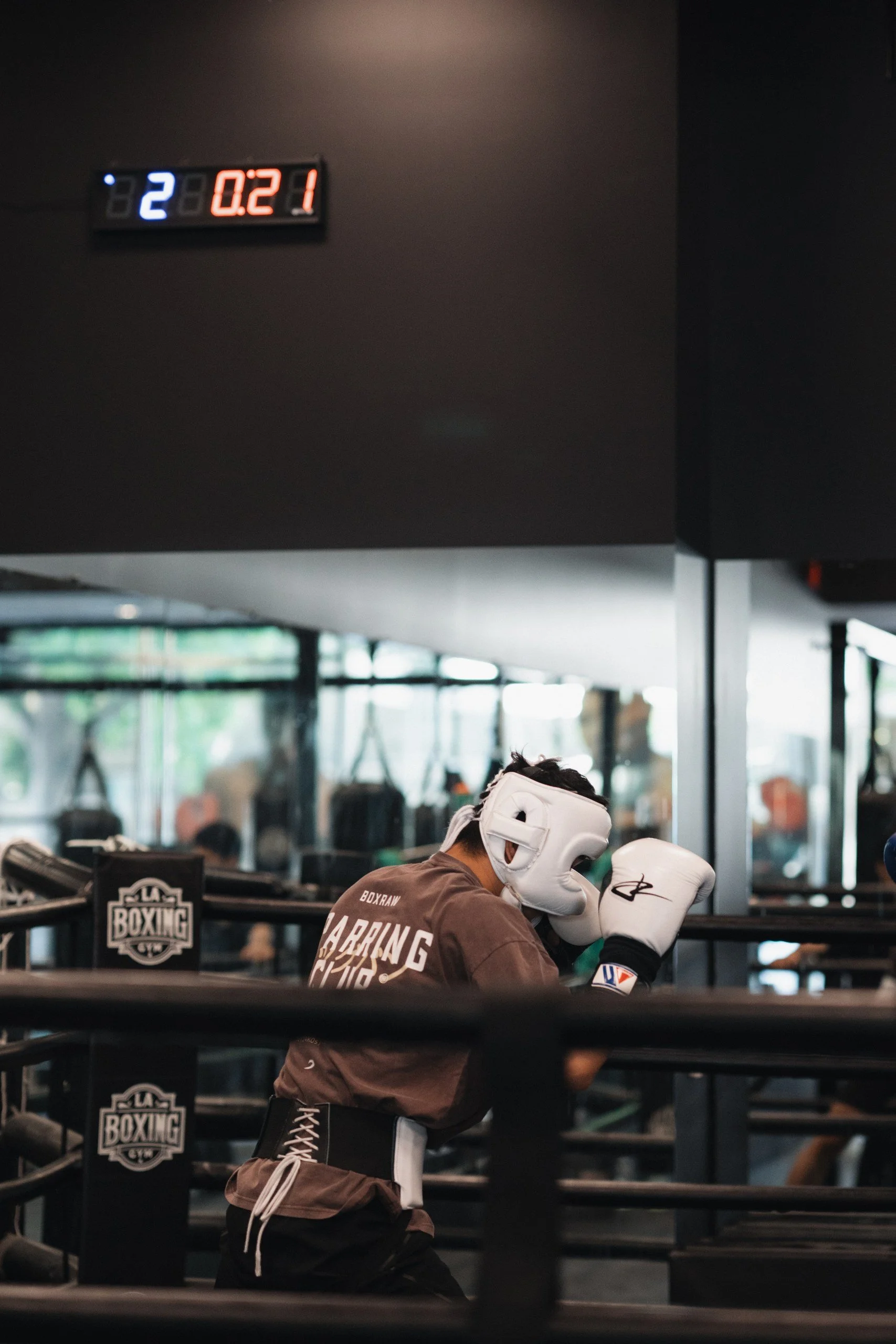 A boxer in training wearing a white headgear and boxing gloves, practicing in a boxing gym with a digital timer displaying 2:21 at the top left, and the gym's boxing logo on the ring post.