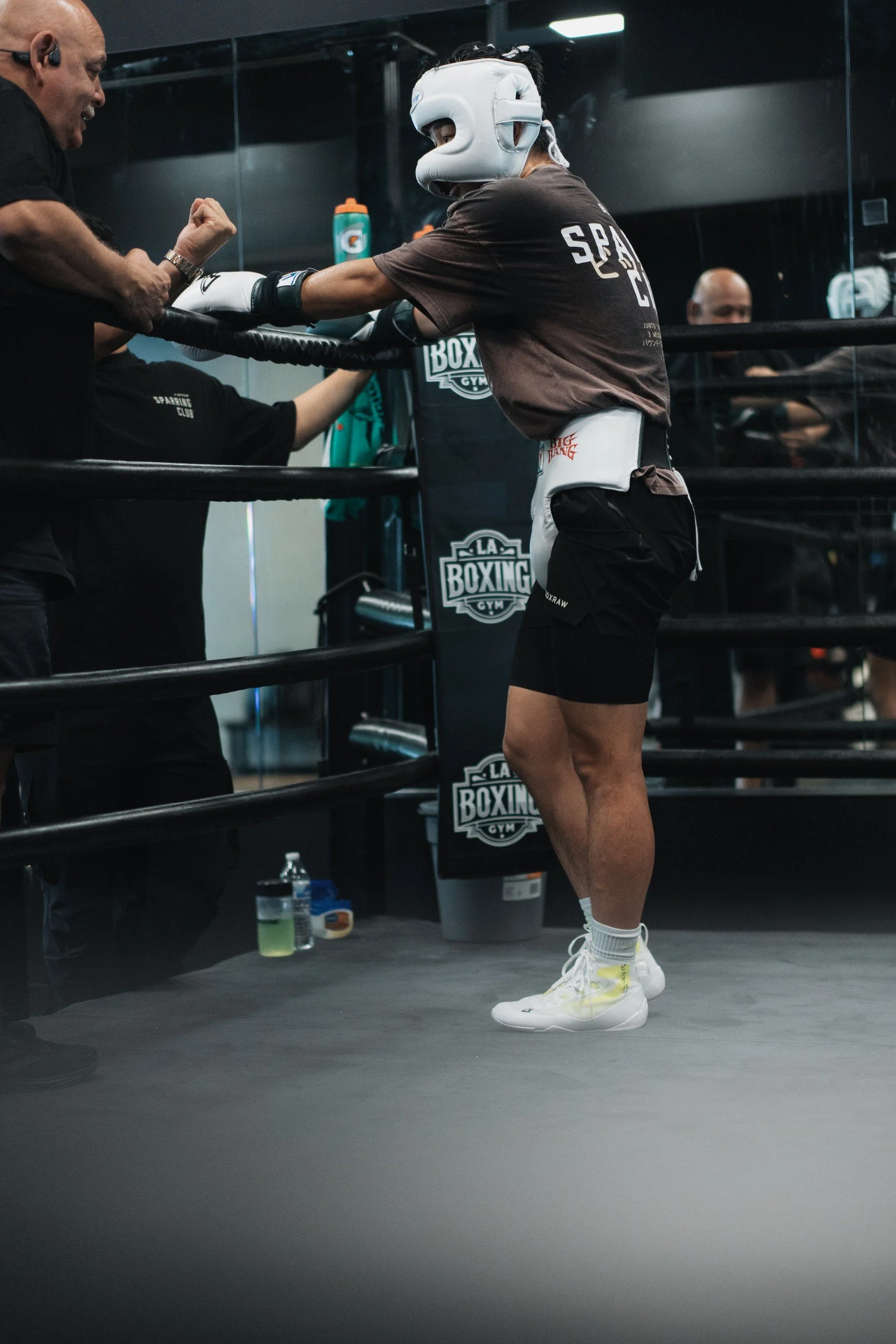 A female boxer wearing protective headgear and boxing gloves in a gym, engaging in a training session with a trainer inside a boxing ring.