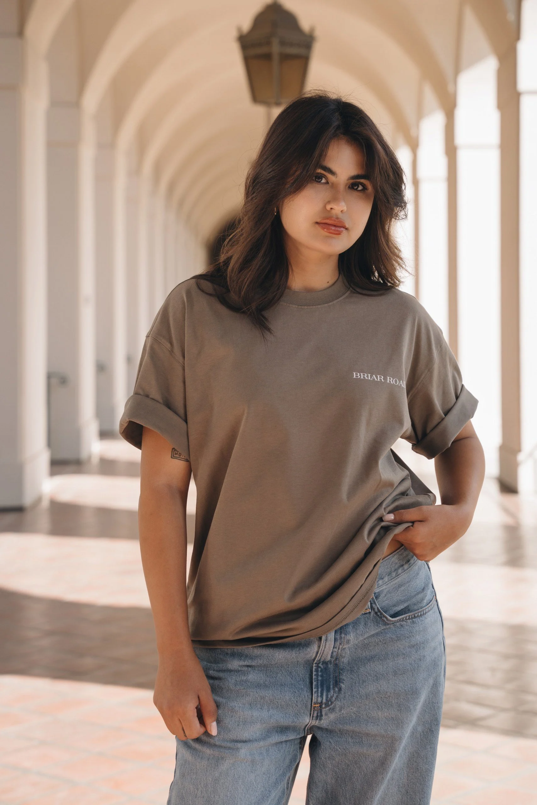 A young woman with dark, shoulder-length hair and light brown skin standing in a covered outdoor walkway with arches and lantern lighting. She is wearing a taupe T-shirt with writing on it and light blue jeans, with one hand in her pocket.