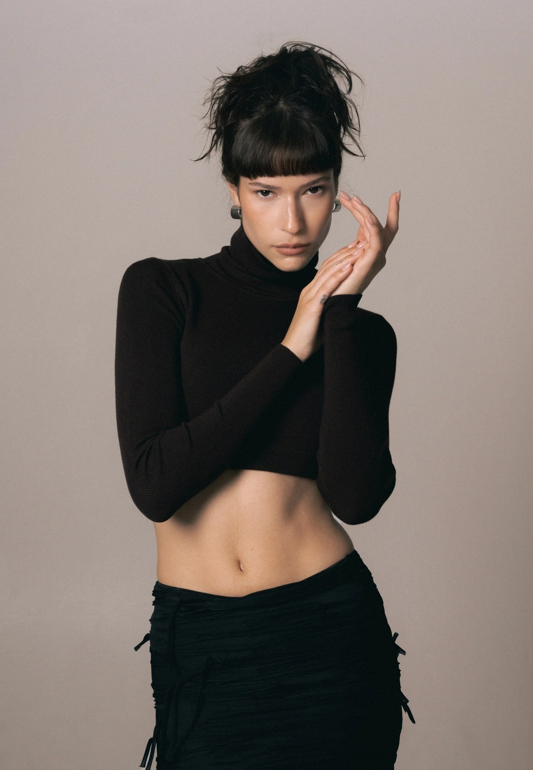 A woman with dark hair styled in a messy updo, wearing a black turtleneck, long black skirt, and earrings, posing against a plain gray background.