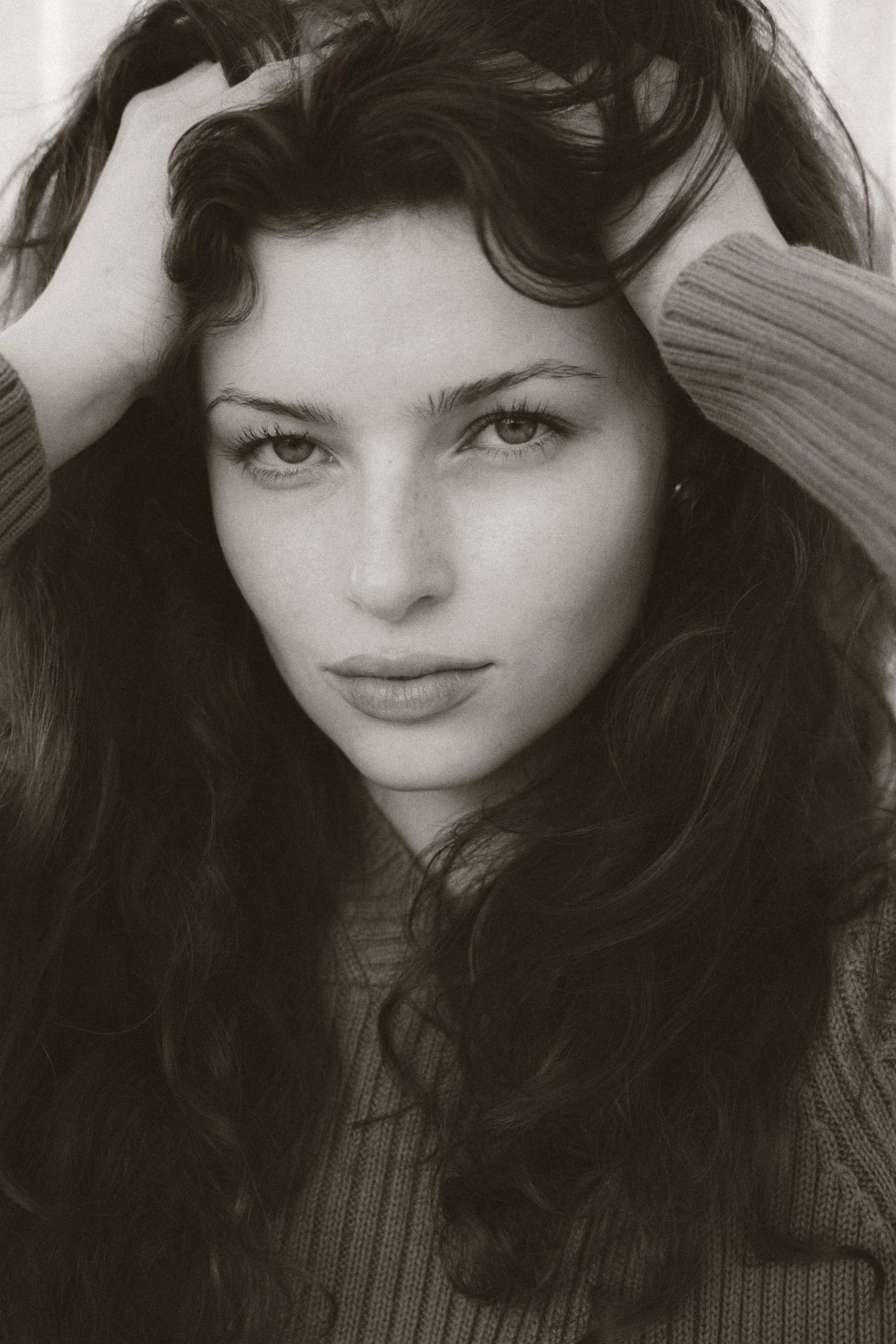 A young woman with long, curly hair, wearing a sweater, holding her hair back with both hands and looking directly at the camera, in black and white.
