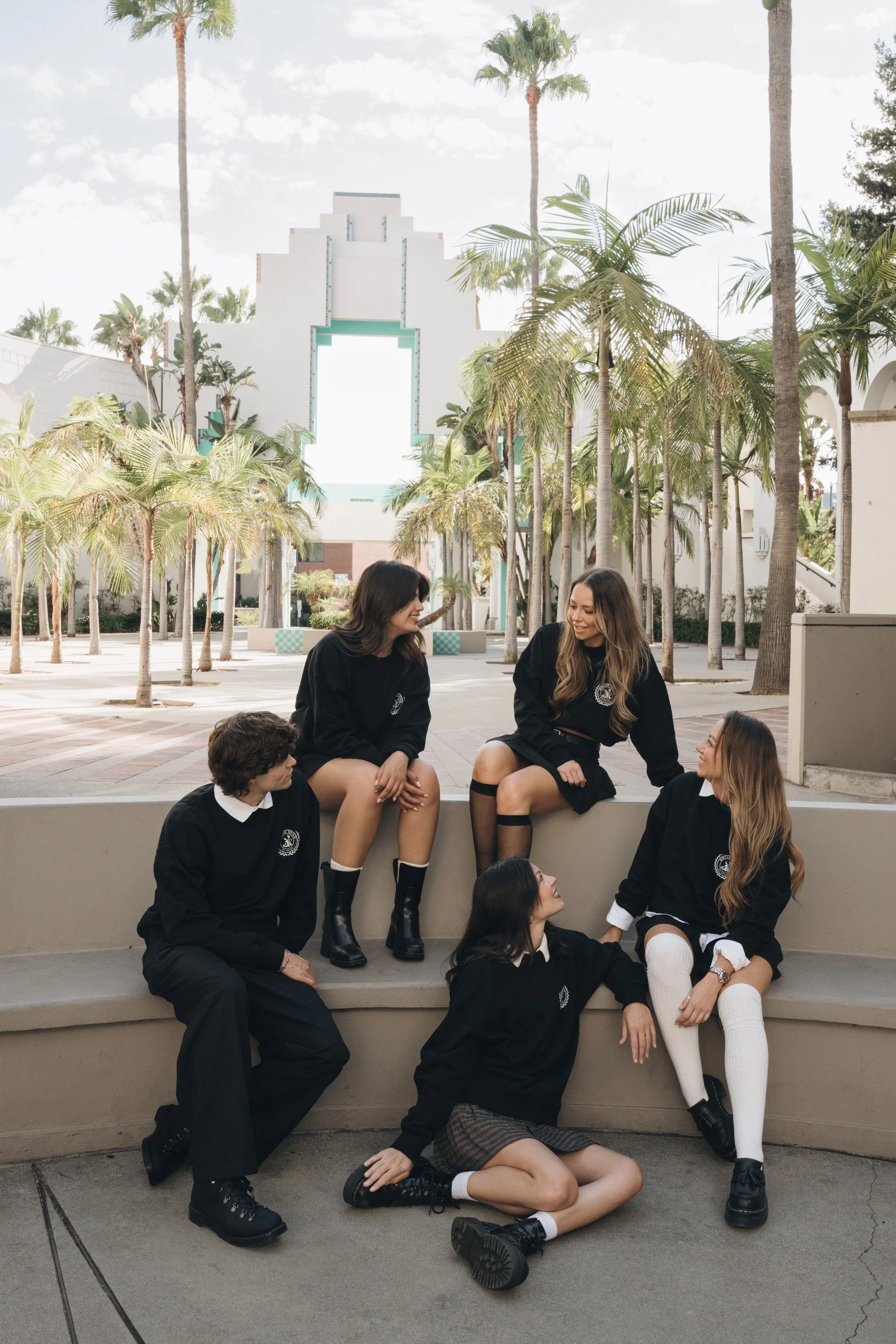 Group of young women dressed in school uniforms sitting and standing on a step outside near palm trees with a white Art Deco building in the background.