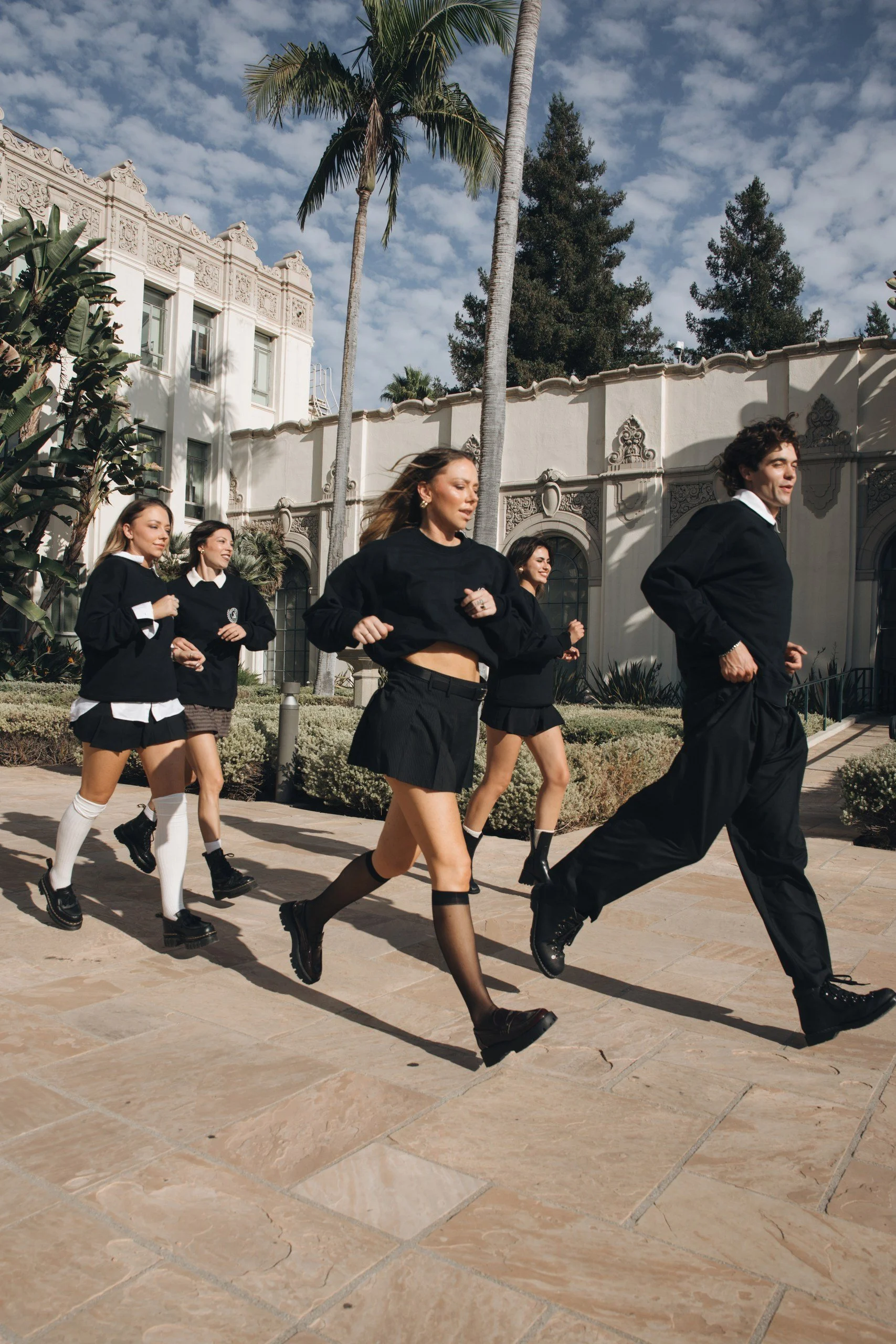 A group of young people dressed in black and white school uniforms, walking outdoors on a sunny day, with tall palm trees, a white building with ornate details, and a partly cloudy sky in the background.