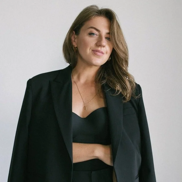 A woman with long brown hair in a blazer and crop top, standing against a plain wall, looking at the camera with a slight smile.