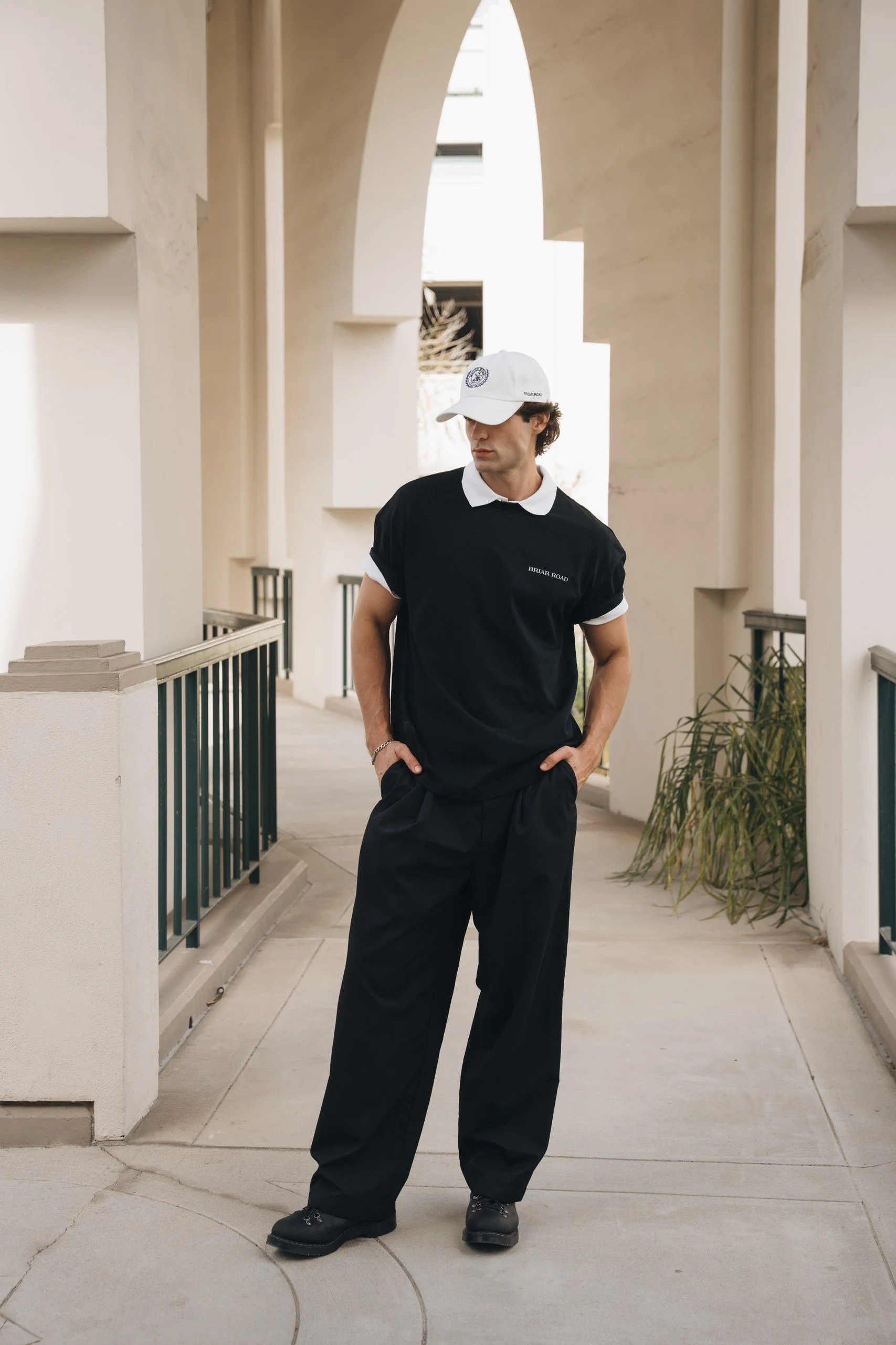 A young man dressed in black clothes, including a t-shirt with 'Briar Road' text, black wide-leg pants, and black shoes, is standing outdoors under an architectural archway with beige walls and plants on the side. He is wearing a white cap with a log