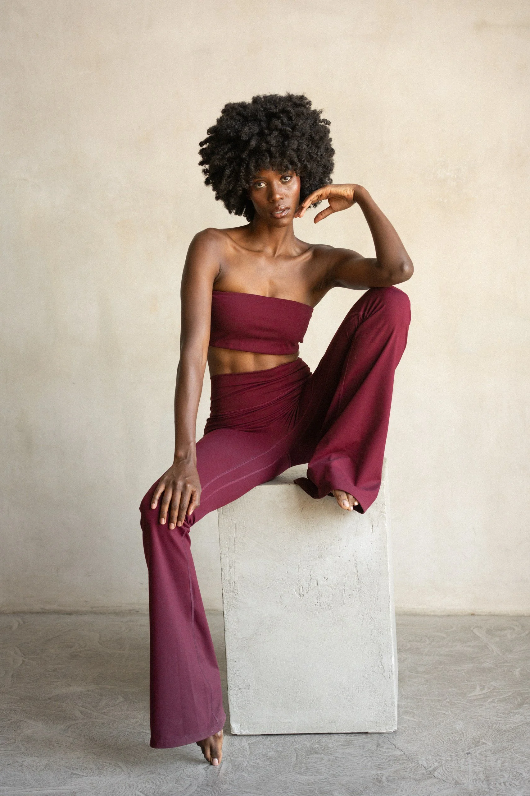A woman with natural curly hair seated on a white block against a plain beige wall. She is wearing a burgundy tube top and matching wide-leg pants, with one leg bent and resting on the block and her hand resting on her thigh, looking at the camera wi