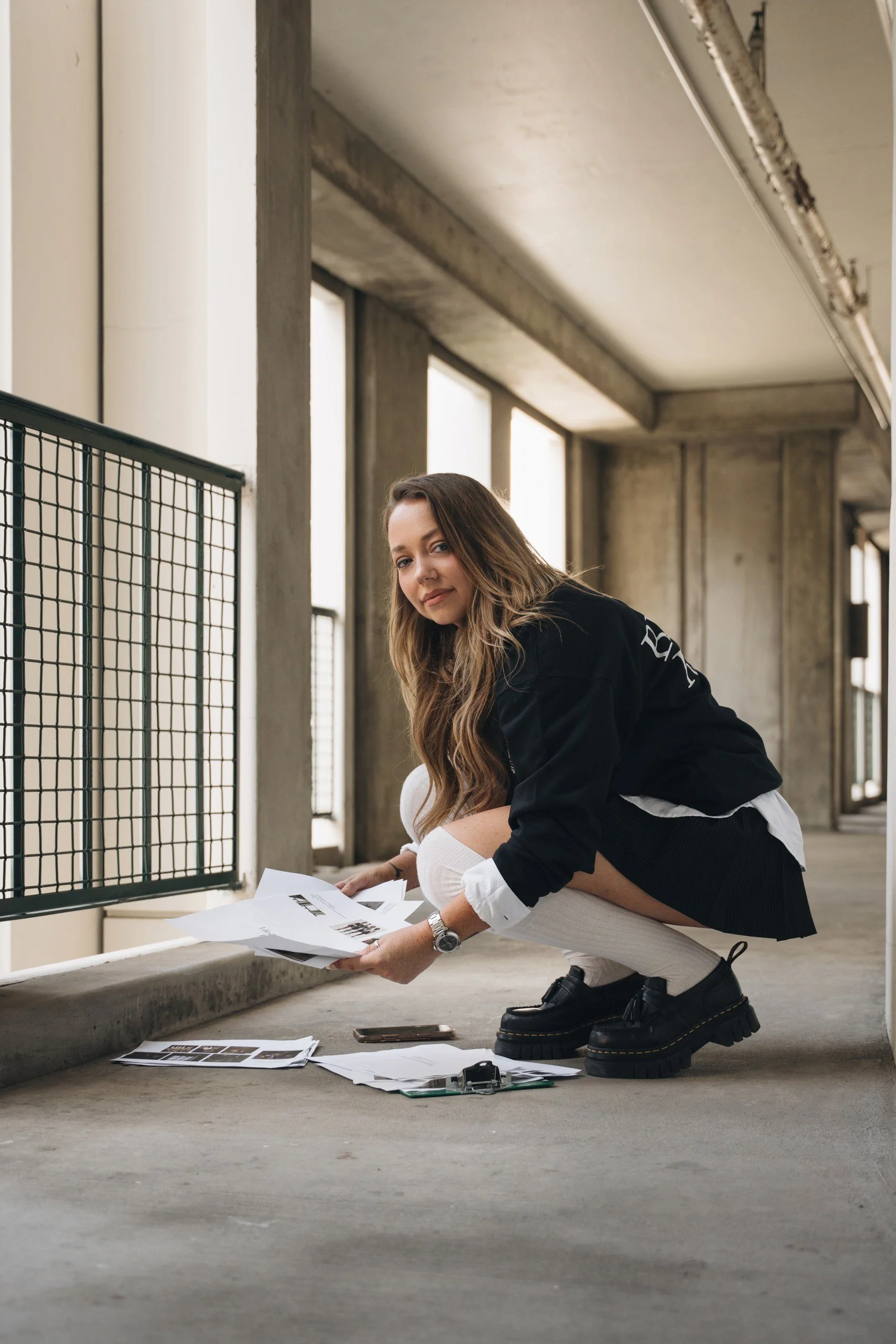 A woman squatting in an industrial-style building with large windows, holding papers and looking at the camera. She is wearing black platform shoes, a black jacket, white socks, and has long wavy hair. There are documents and a smartphone on the grou