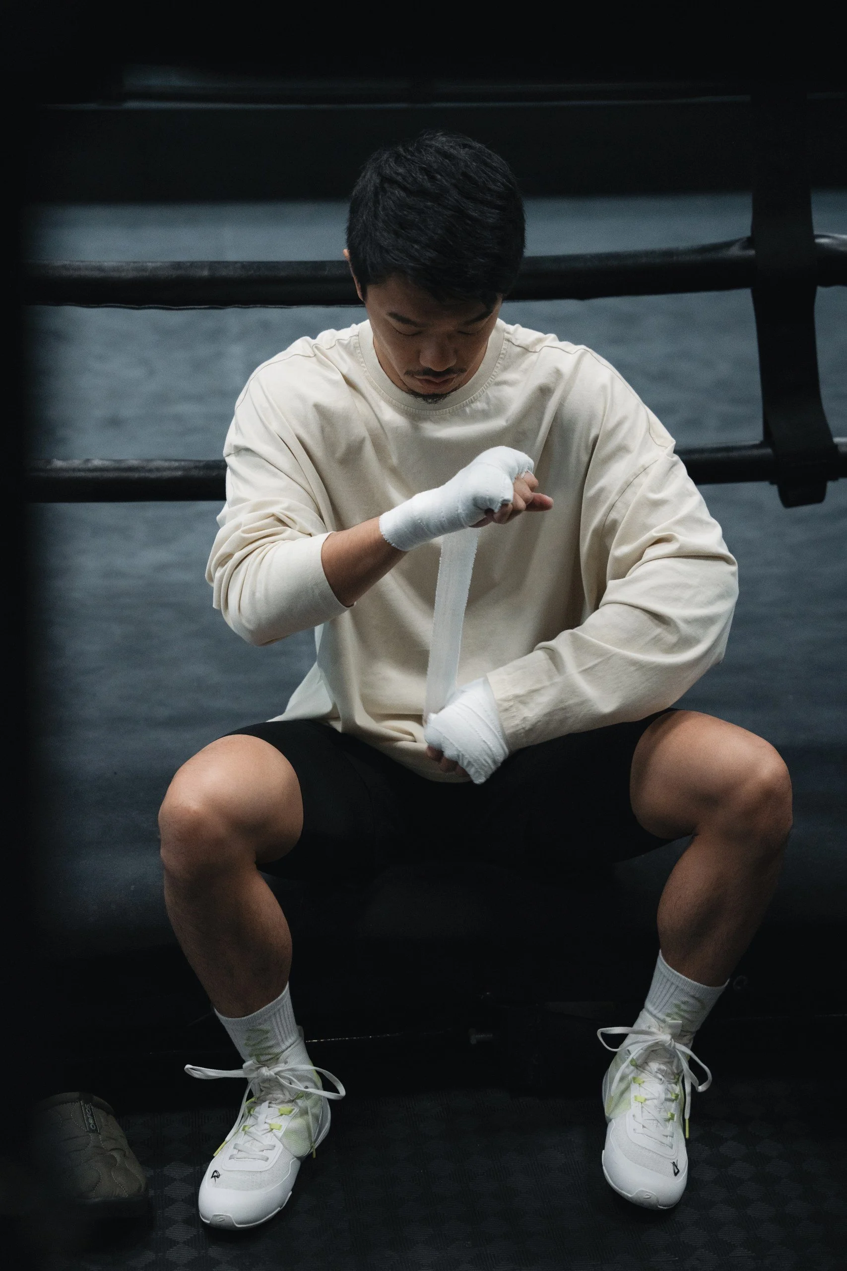 A male athlete sitting down in a gym, wrapping his hands with white hand wraps, wearing a cream-colored long-sleeve shirt, black shorts, and white athletic shoes, with black boxing or MMA gloves placed beside him on the floor.