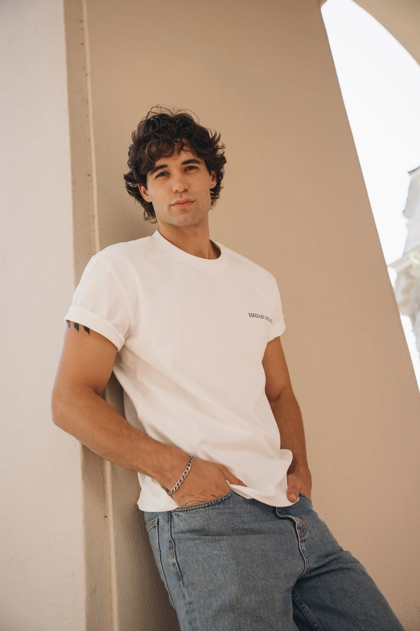 A young man with curly dark hair and light skin leaning against a beige wall, wearing a white t-shirt and blue jeans.