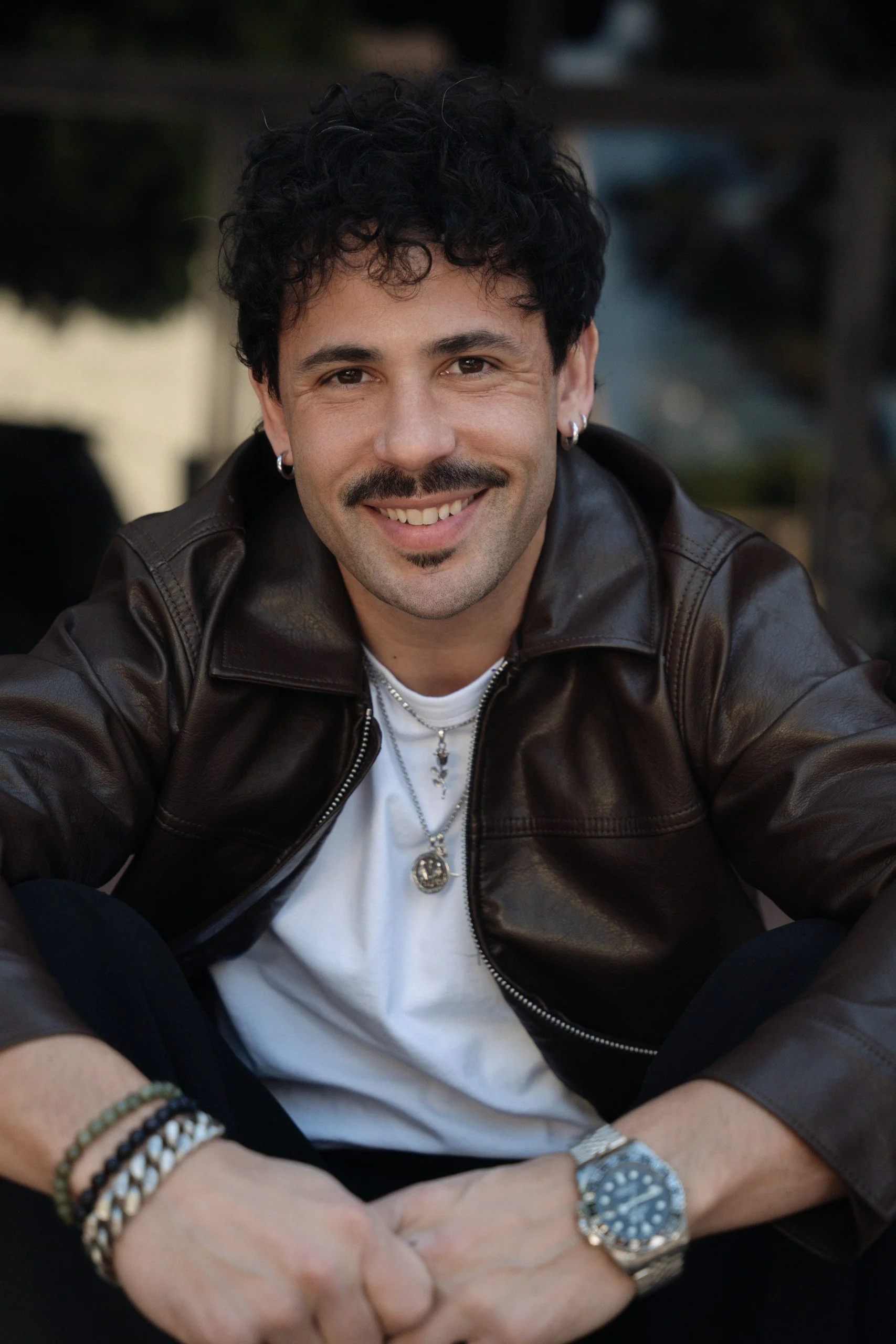 A young man with curly black hair, wearing earrings, smiling, dressed in a brown leather jacket over a white shirt, with jewelry including bracelets, necklaces, a watch, and rings, sitting outdoors.