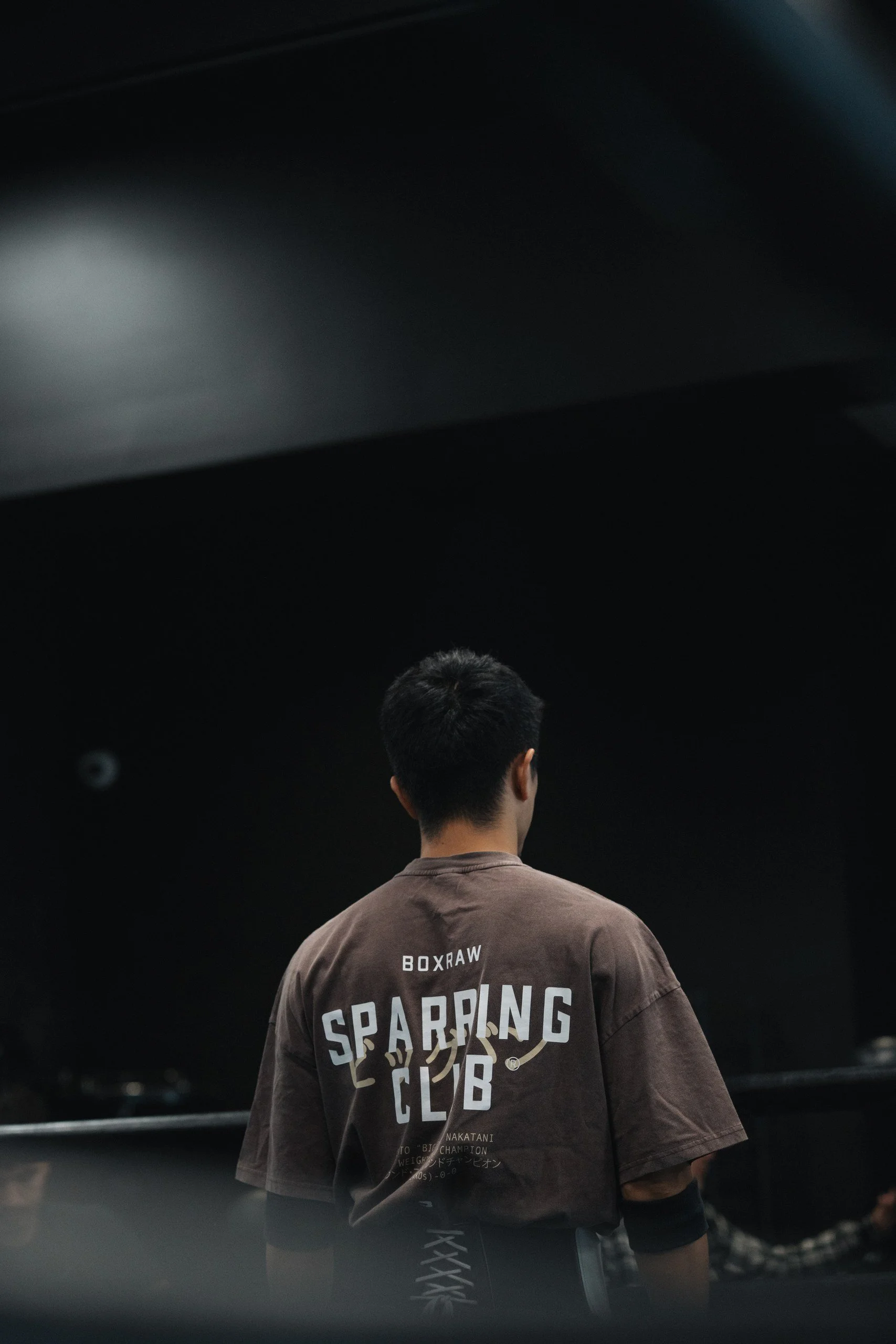 The back of a man wearing a brown T-shirt with the text 'Box Raw' and 'Scraping Club' in large letters, standing in a dark room, possibly a gym or training environment, with his head slightly turned and short dark hair.