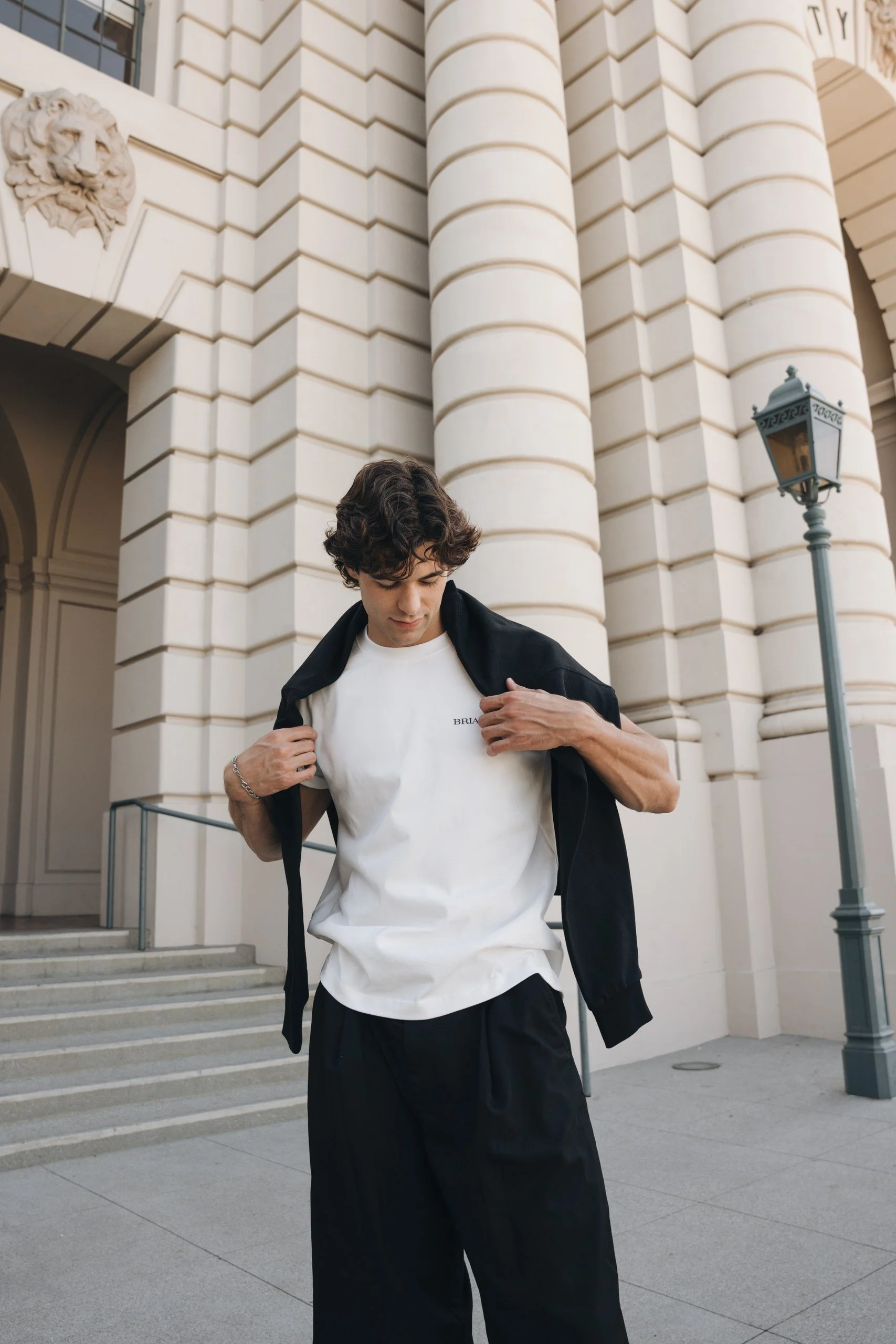 A young man with curly dark hair and light skin standing outside a grand building with large white columns and detailed architecture. He's wearing a white T-shirt, black pants, and a black jacket, which he is adjusting.