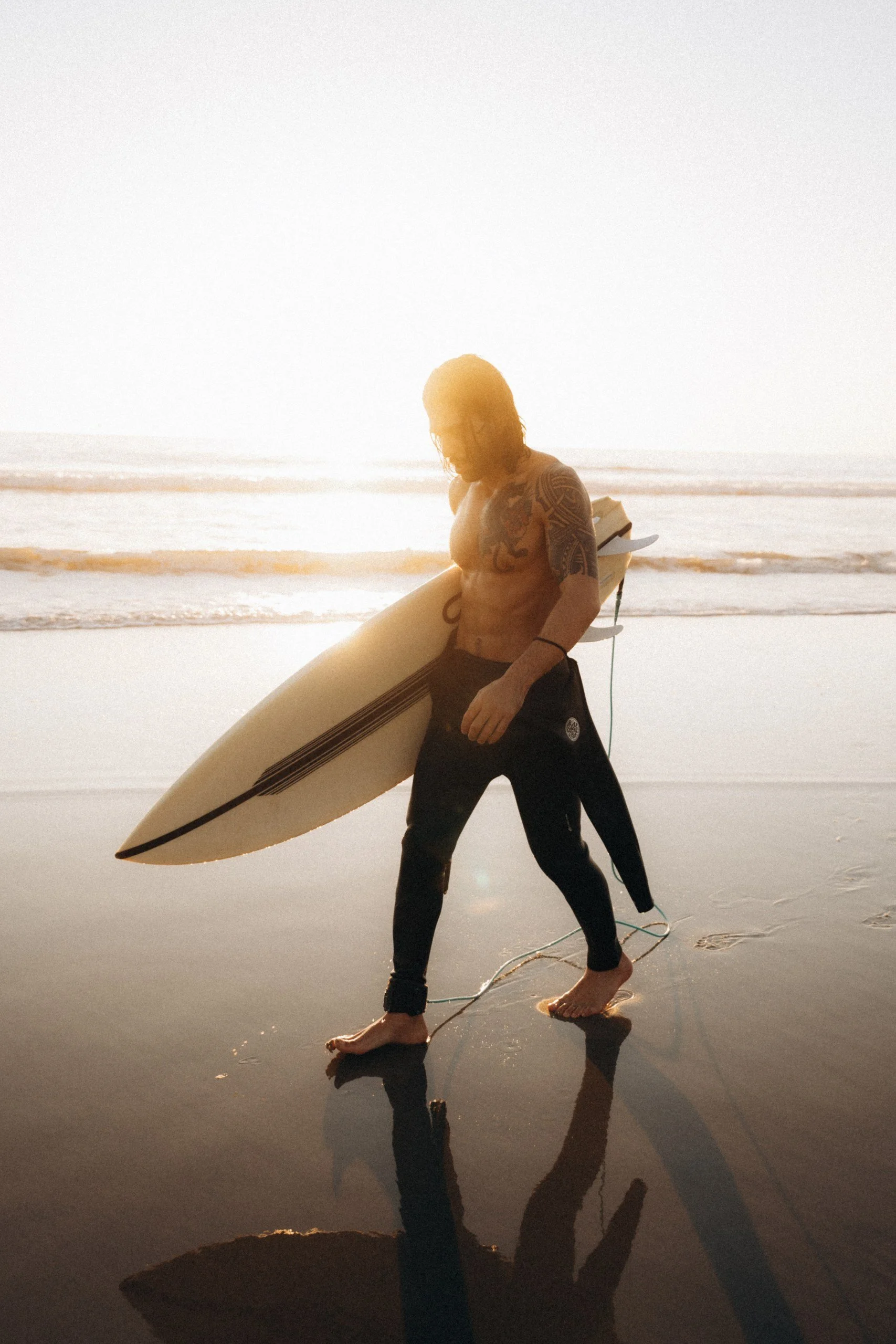 A shirtless man with tattoos on his arms and chest carrying a surfboard at the beach during sunset or sunrise.