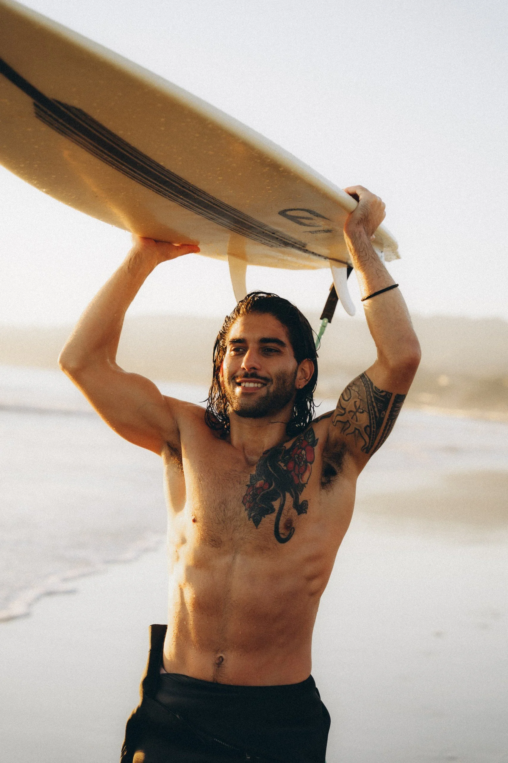 A shirtless man with tattoos on his chest and arm, holding a surfboard above his head at the beach during sunset.