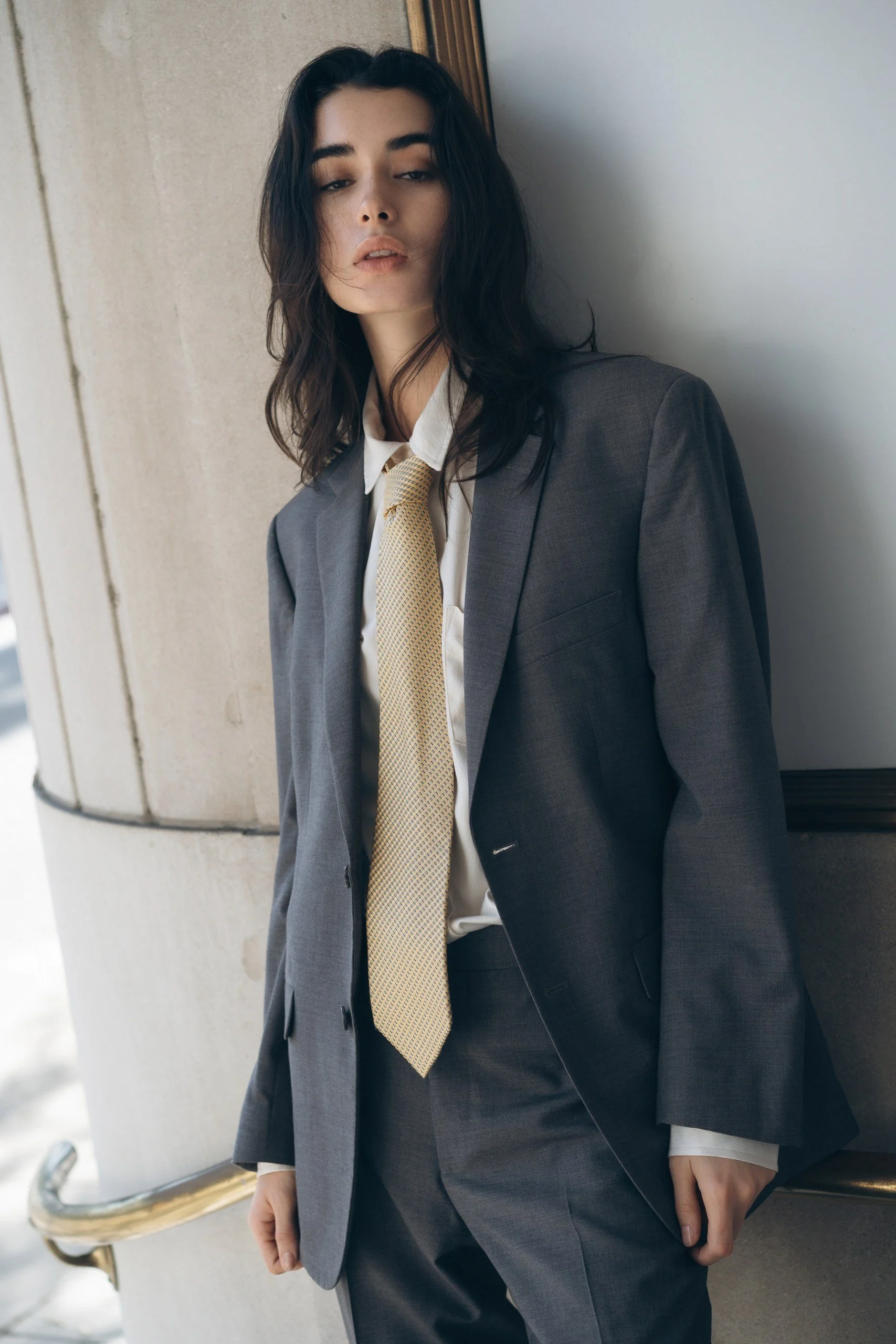 A woman with dark, wavy hair in a gray suit with a yellow tie, leaning against a wall with a neutral expression.