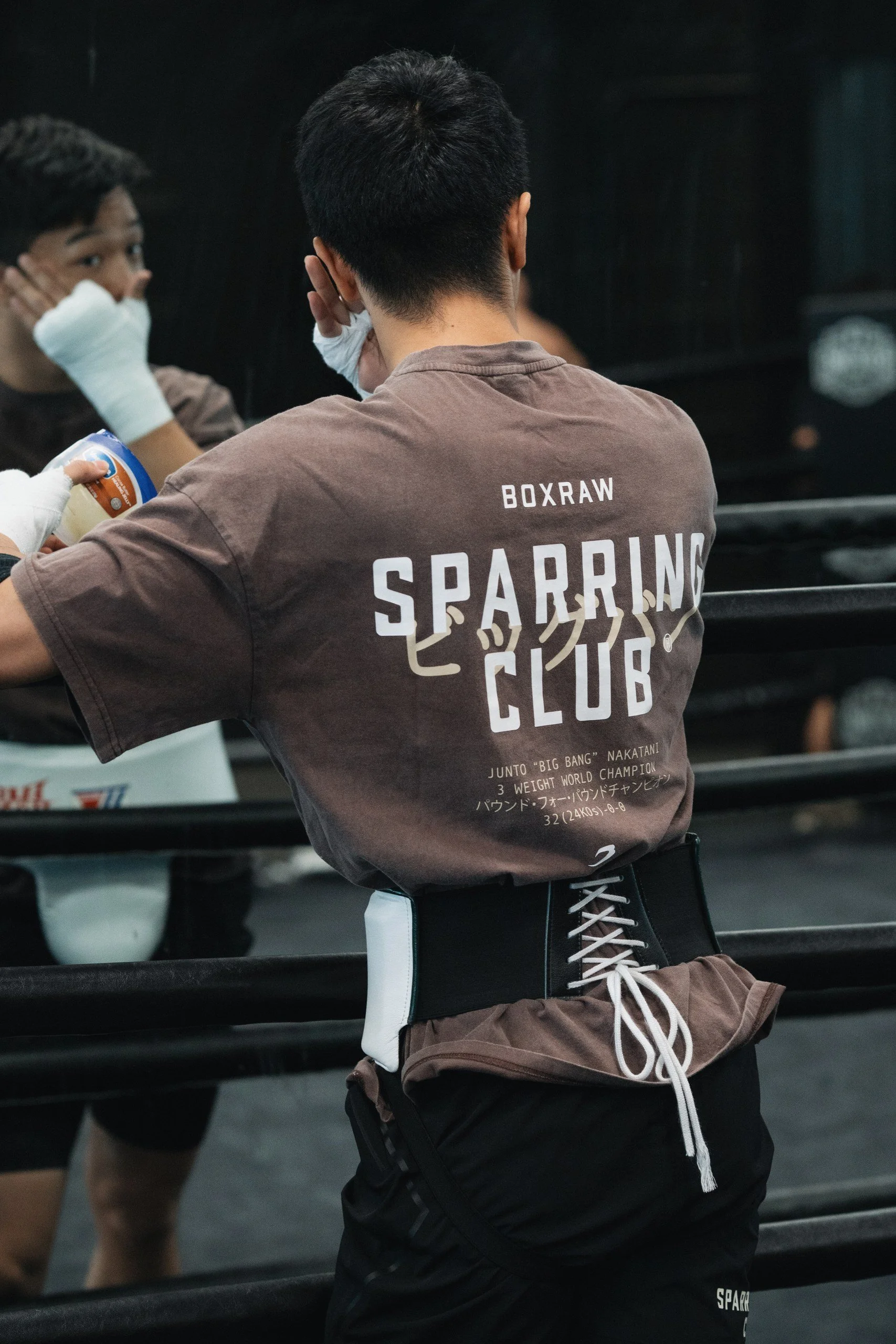 Two men in a gym, one wearing a gray T-shirt with white lettering that reads 'SPARRING CLUB' and black shorts, and the other in a brown shirt and black shorts, both wearing boxing gloves and standing near a boxing ring.