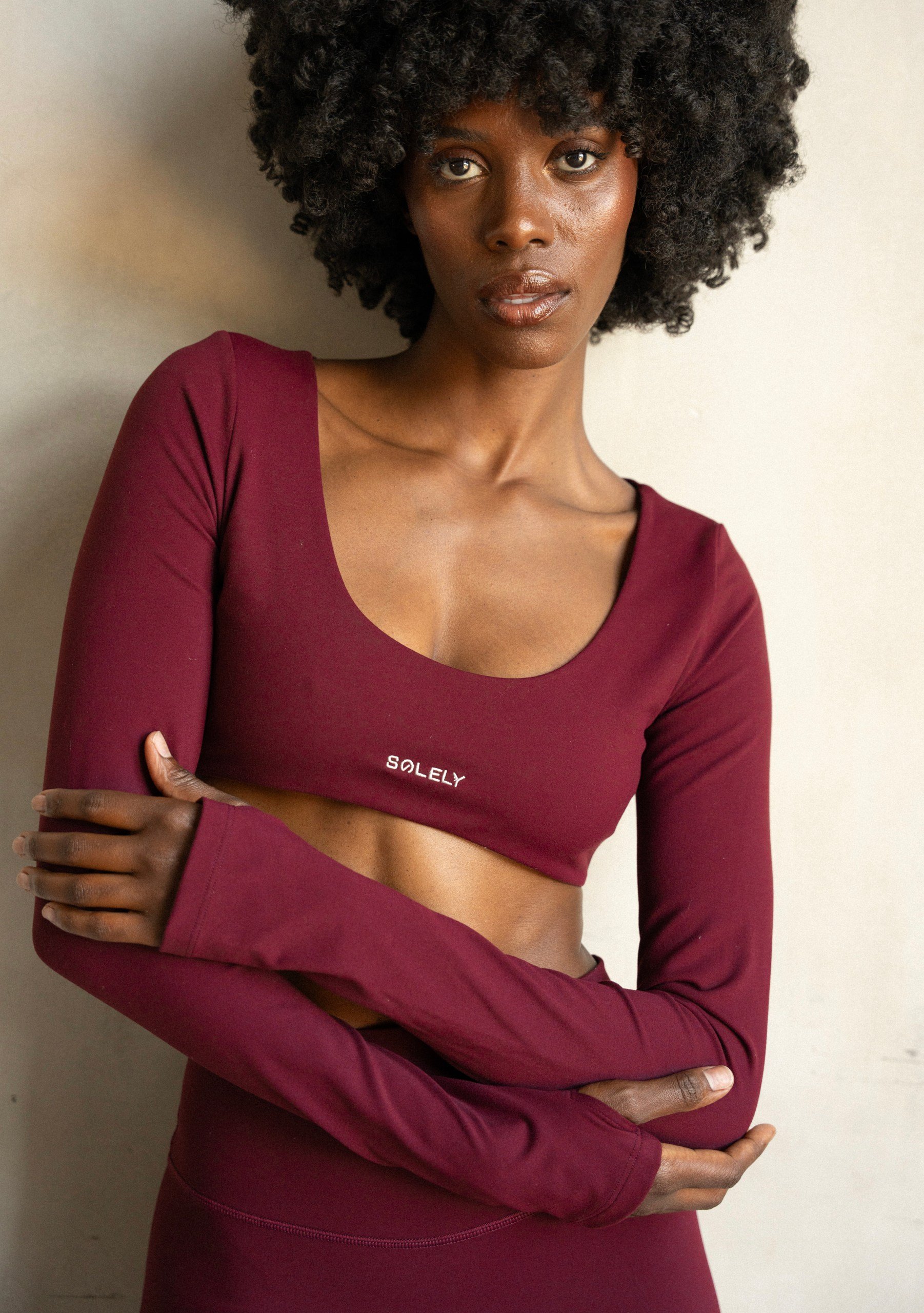A woman with dark skin and curly black hair wearing a maroon long-sleeve crop top with the word 'SOLELY' written on it, standing against a plain background.