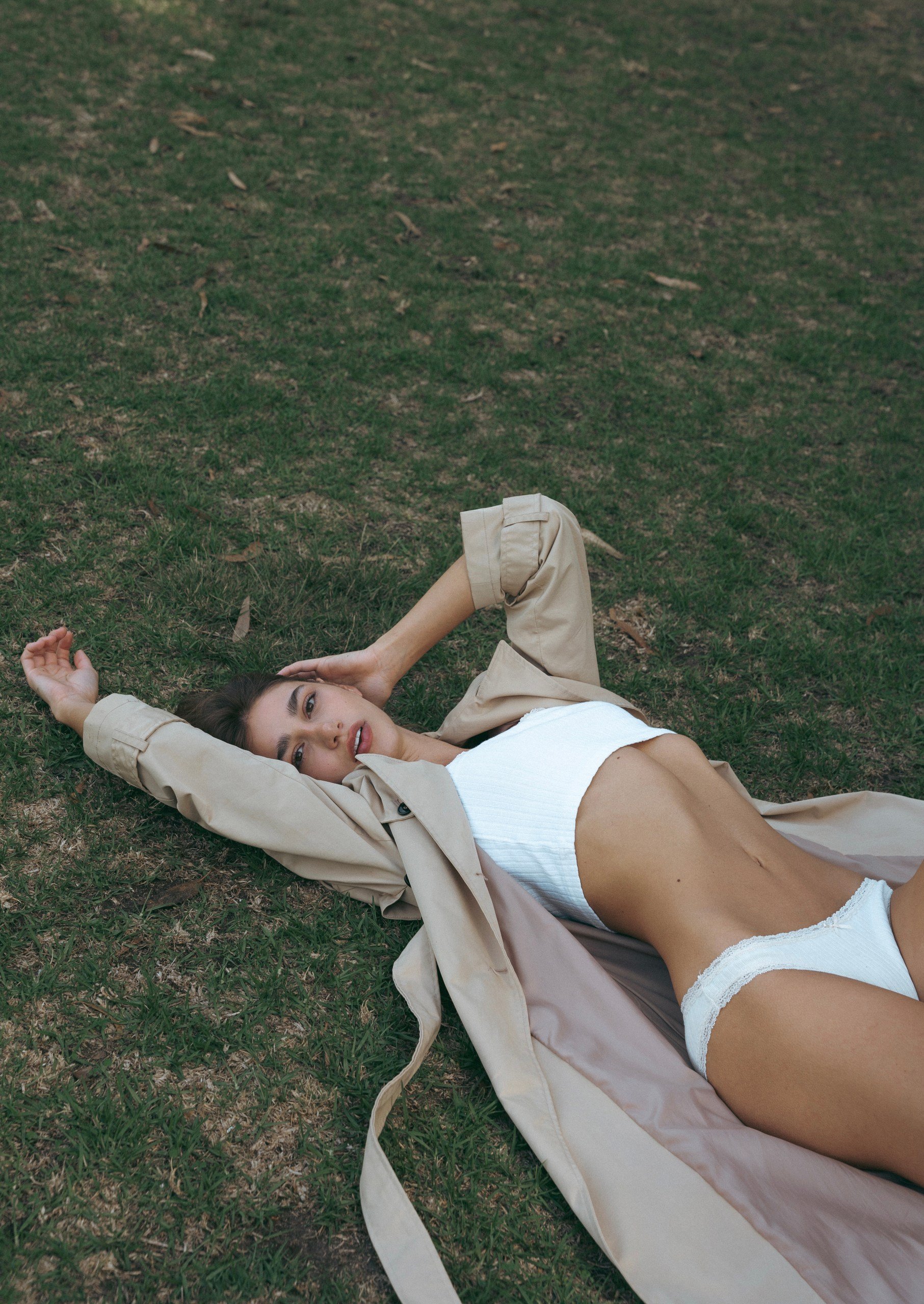 Young woman lying on grass in a relaxed pose, wearing a beige jacket and white lingerie, outdoors during daytime.