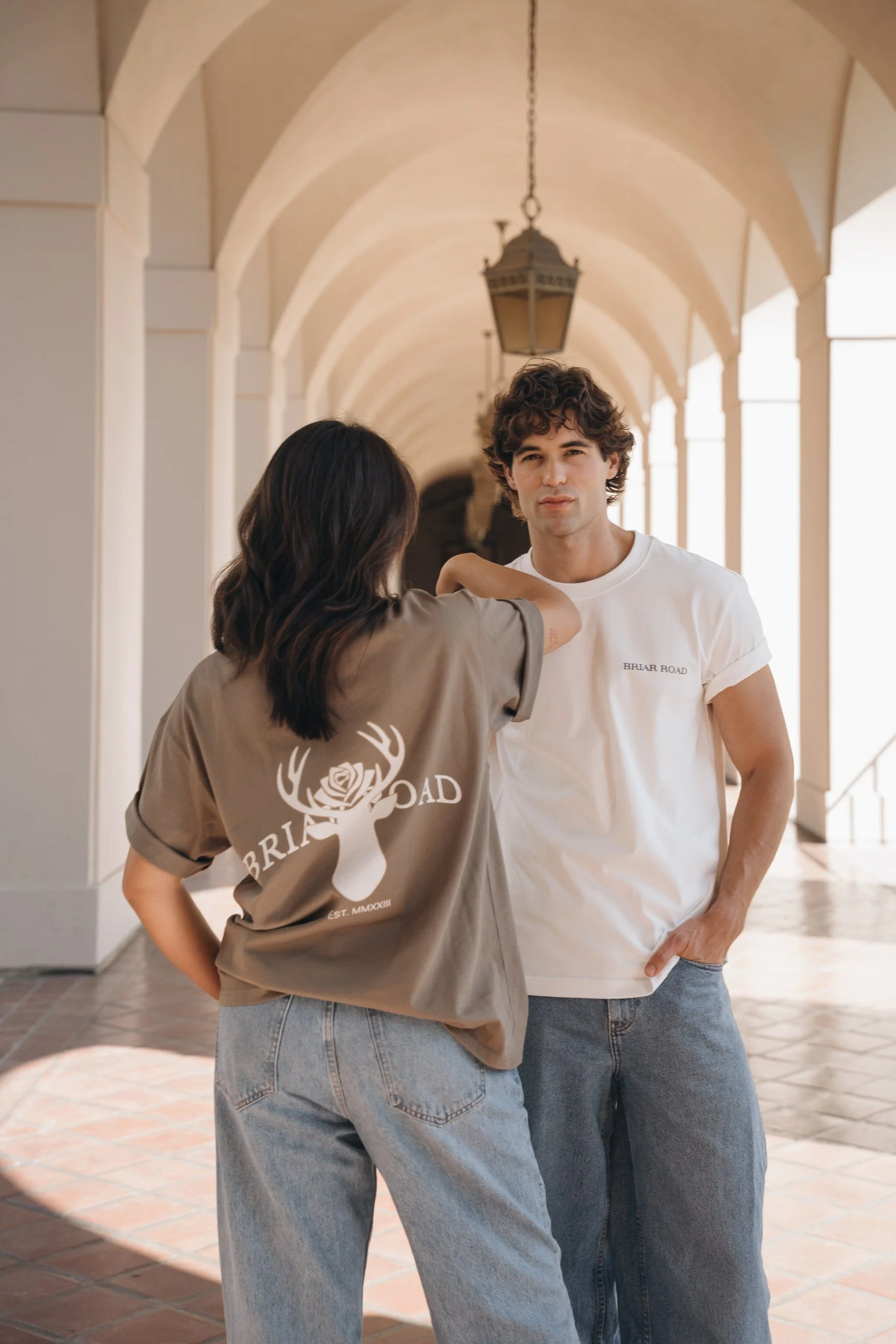 A woman with dark hair is standing with her back to the camera, resting her arm on the shoulder of a man with curly hair. The man is facing forward, and both are wearing casual clothing, with the woman in a beige T-shirt with a printed logo and the m