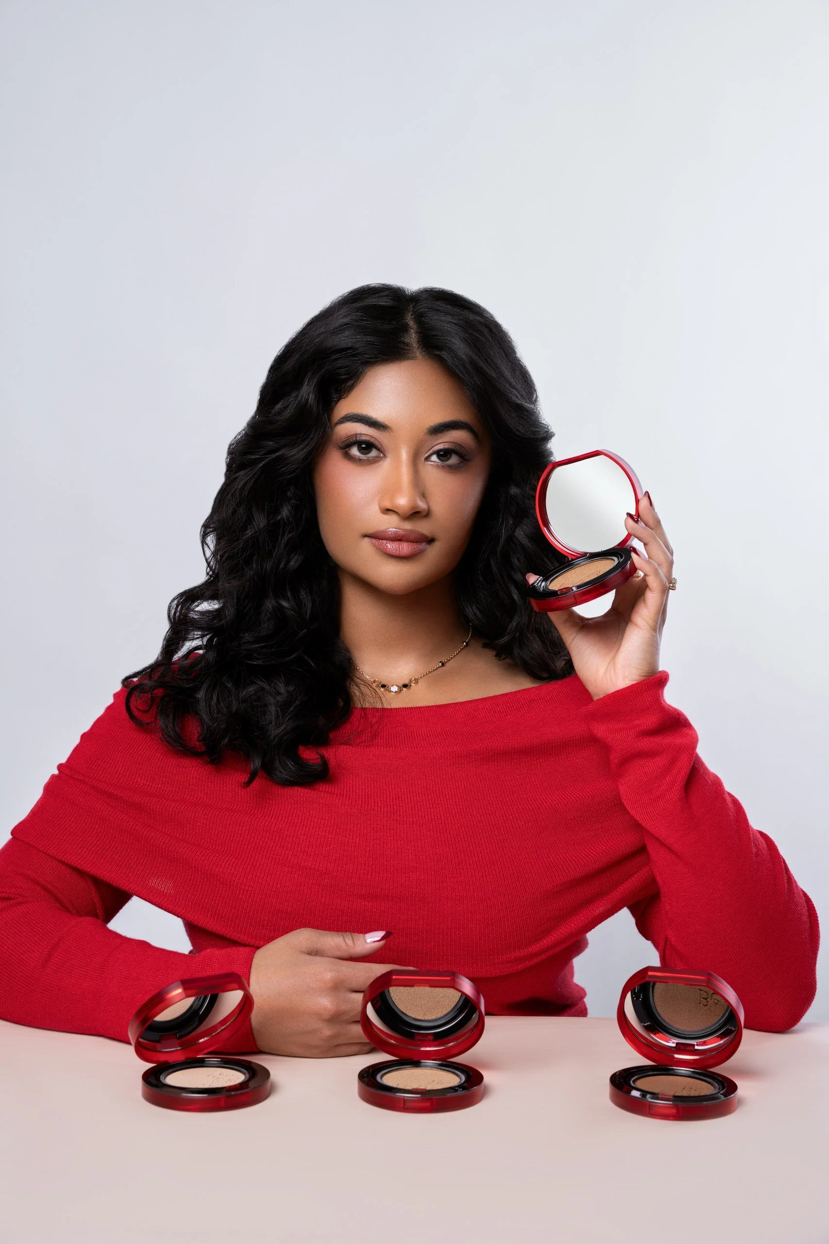 Woman in a red sweater holding an open compact mirror with makeup inside, with other open makeup compacts on a white surface in front of her, against a light gray background.