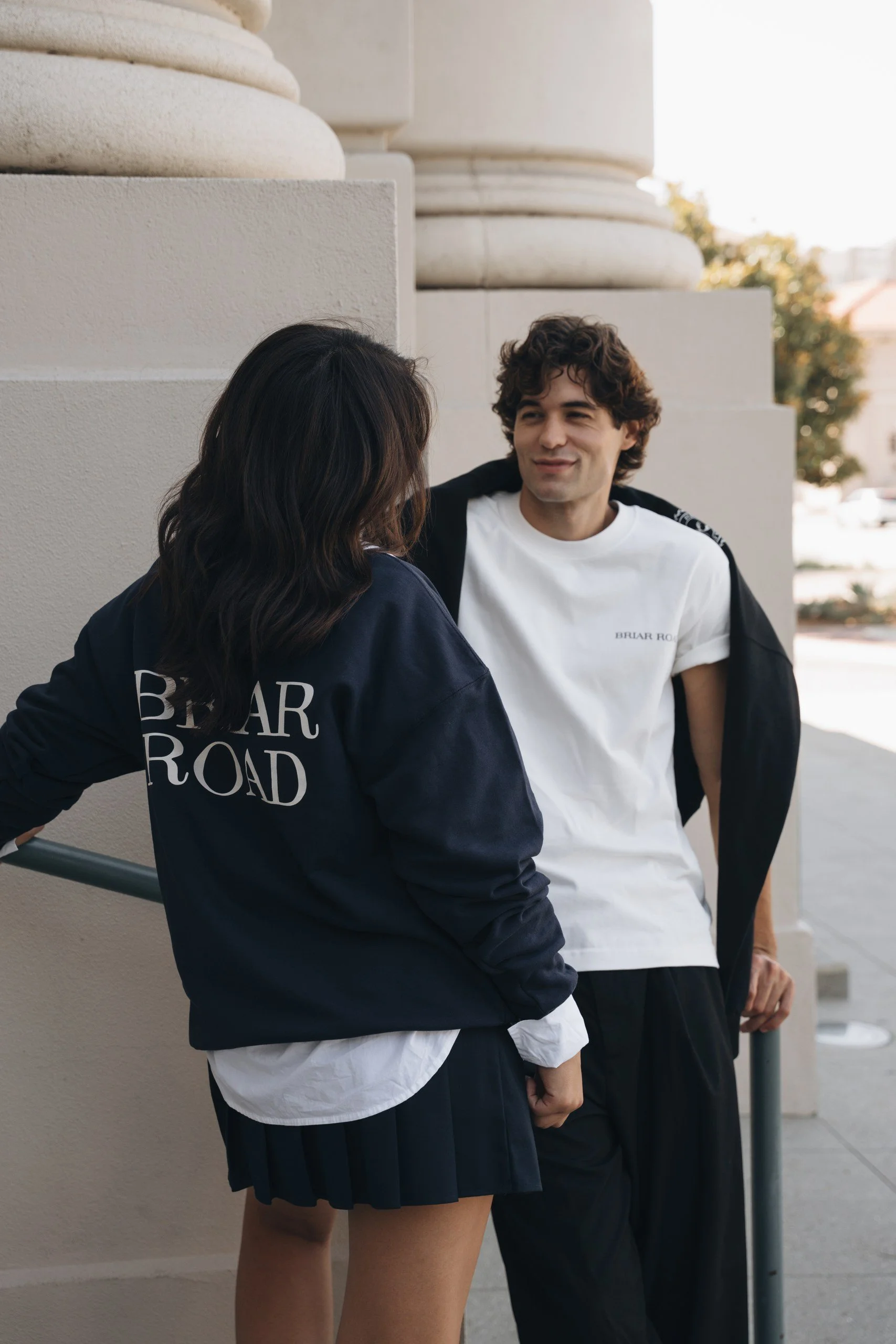 A young woman and a young man are standing outdoors near a building. The woman has dark hair and is wearing a navy jacket with white lettering, while the man has curly brown hair and is wearing a white T-shirt and a black jacket. They are engaged in 