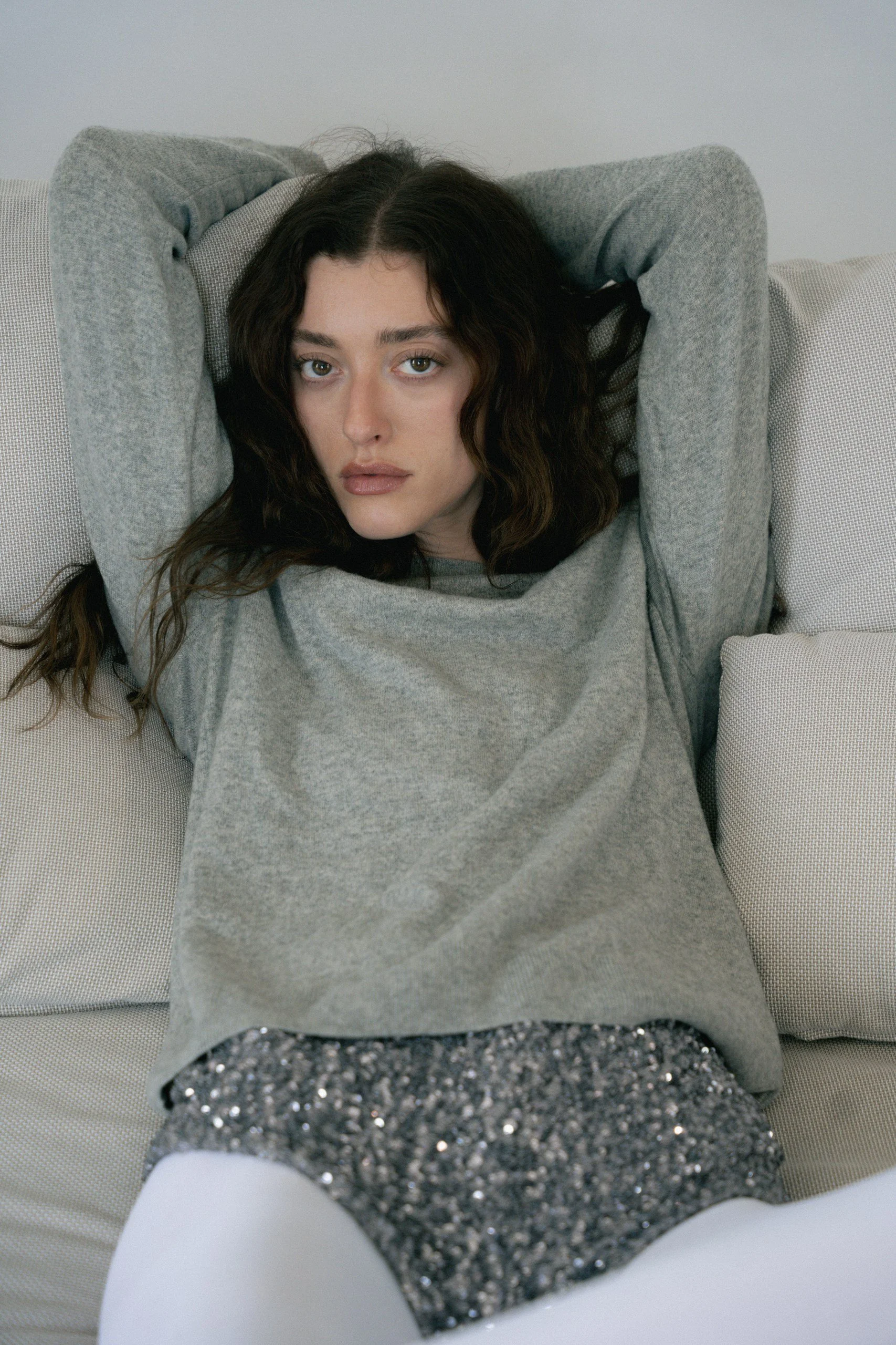 A young woman with dark curly hair and fair skin lying on a couch with her hands behind her head, wearing a gray sweatshirt, a sparkly skirt, and white tights.