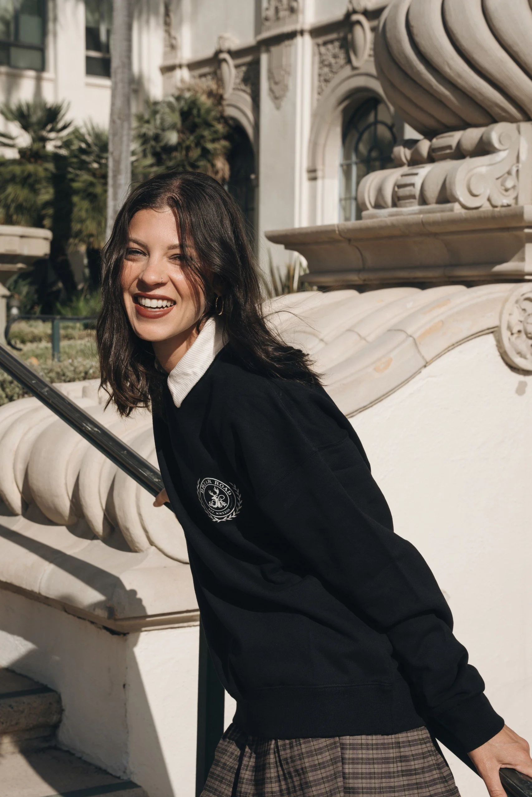 A young woman with dark hair and light skin smiling and sticking out her tongue, standing outdoors in front of a decorative stone structure and plants, wearing a black jacket and a plaid skirt.